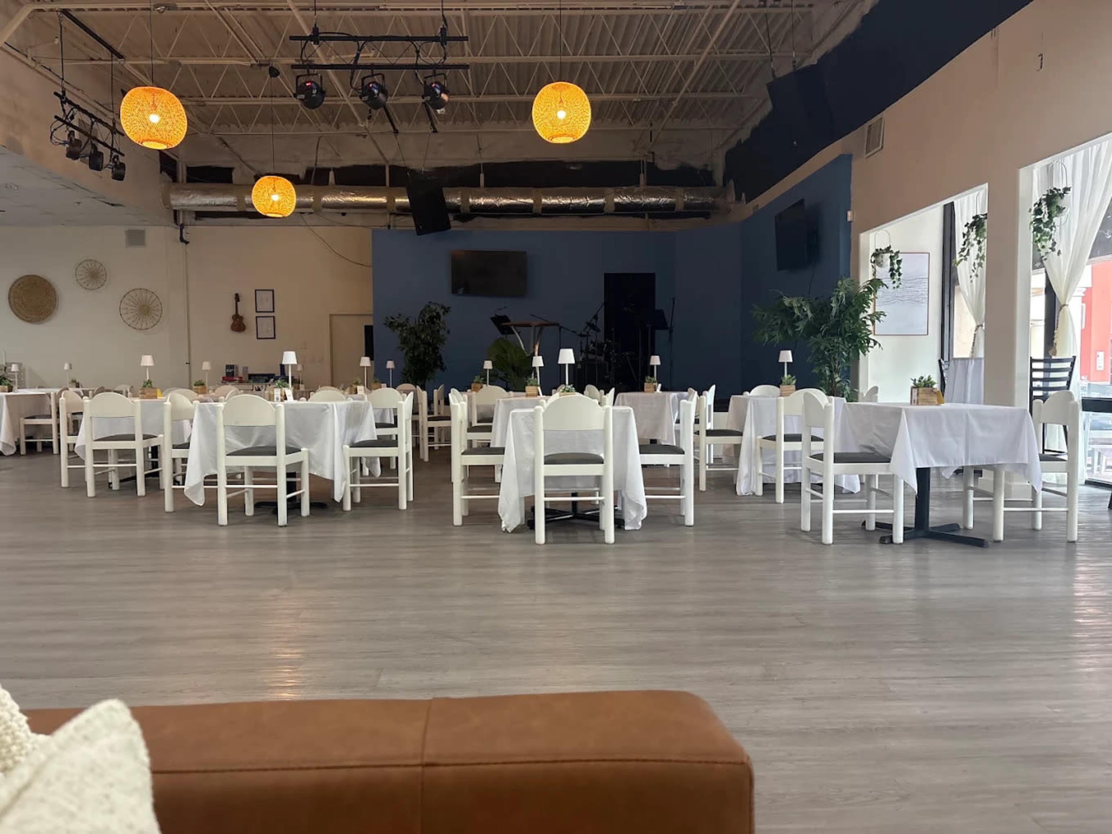 The image shows a spacious restaurant with white tables covered in linens, arranged in a neat layout, and featuring pendant lights hanging from the ceiling.
