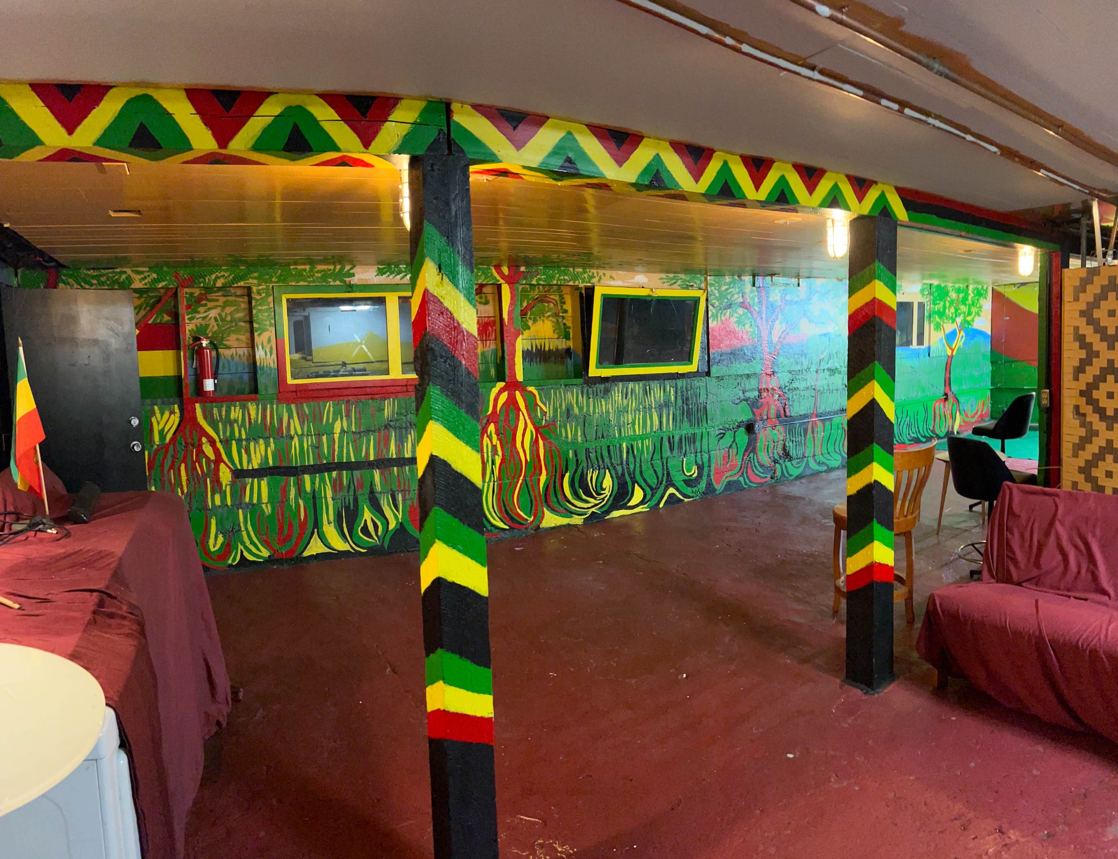 The image shows a colorful room featuring vibrant murals on the walls, with wooden beams painted in a striped pattern, and various items like chairs and flags present in the space.