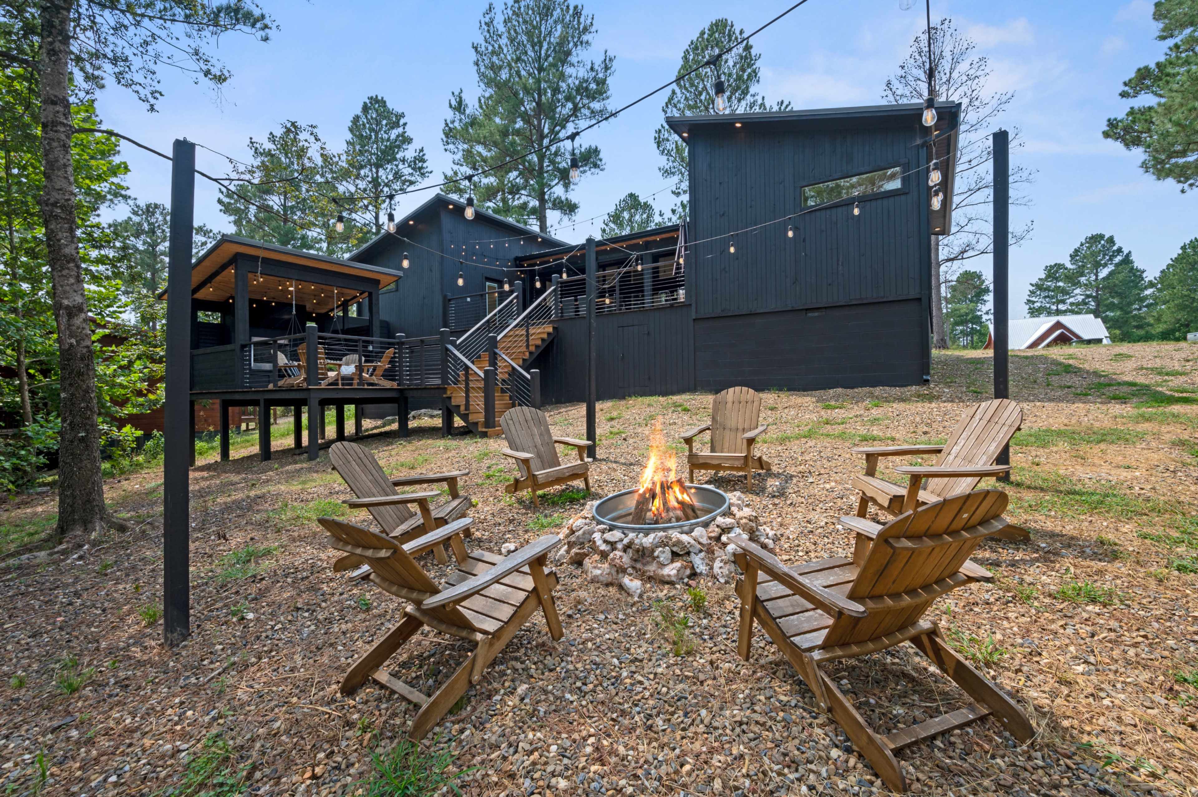 A modern black house is set among trees, featuring a fire pit surrounded by wooden chairs, with string lights overhead and a gravel pathway leading to it.
