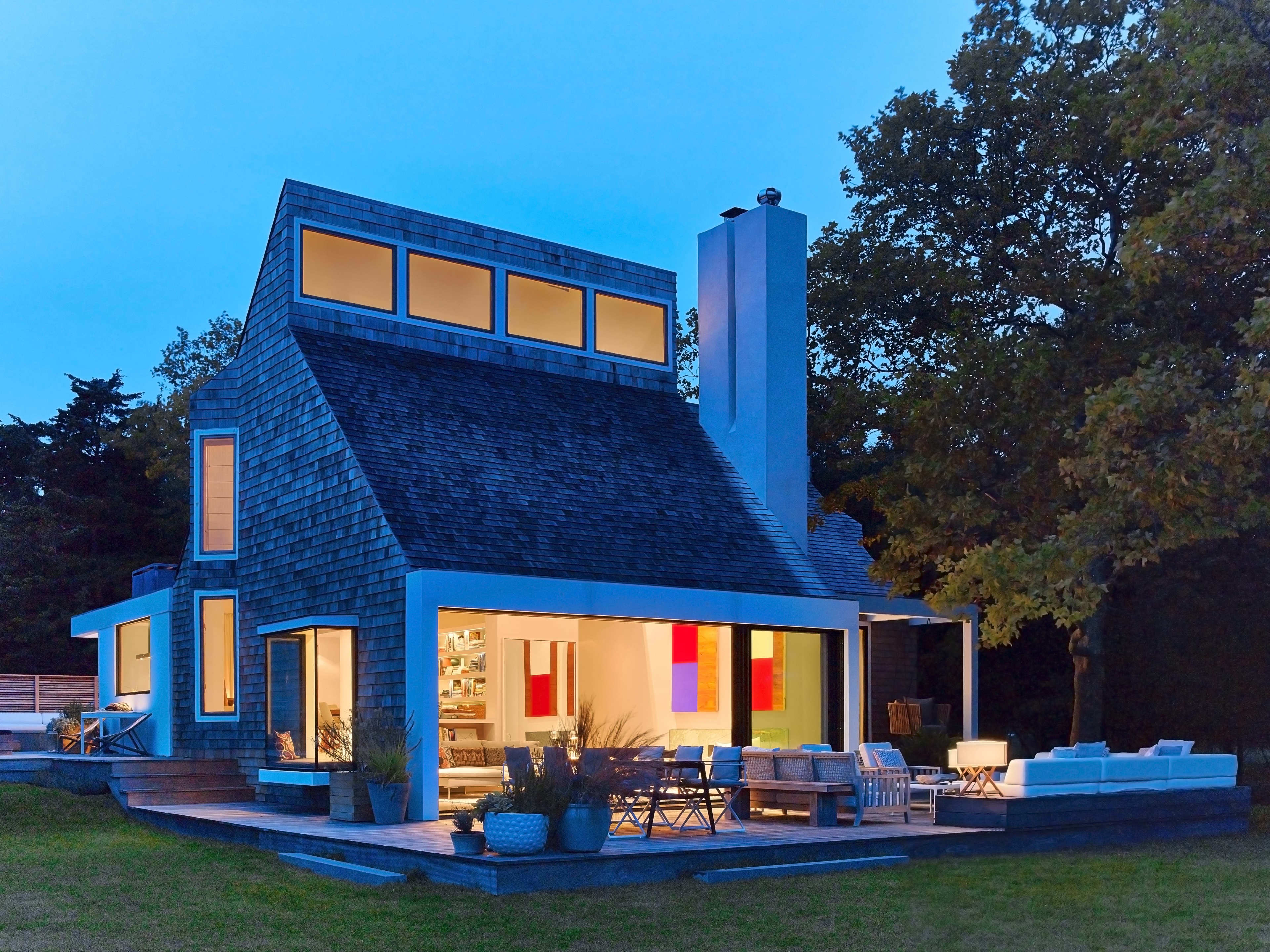 A modern house with large windows and a sloped roof is illuminated in the evening, surrounded by a lawn and outdoor furniture.