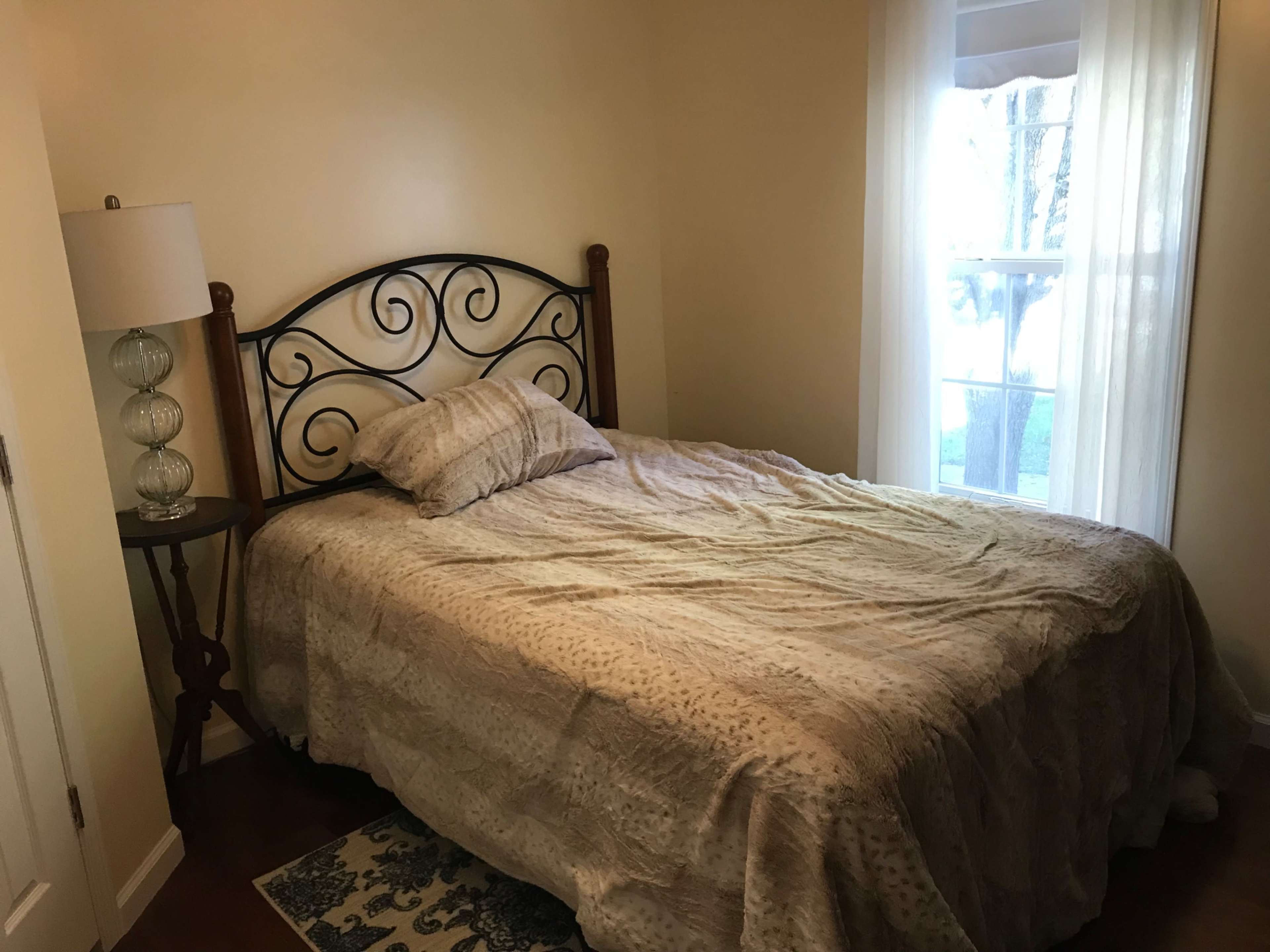 A small bedroom with a bed covered by a patterned comforter, a bedside table with a lamp, and a window letting in natural light.