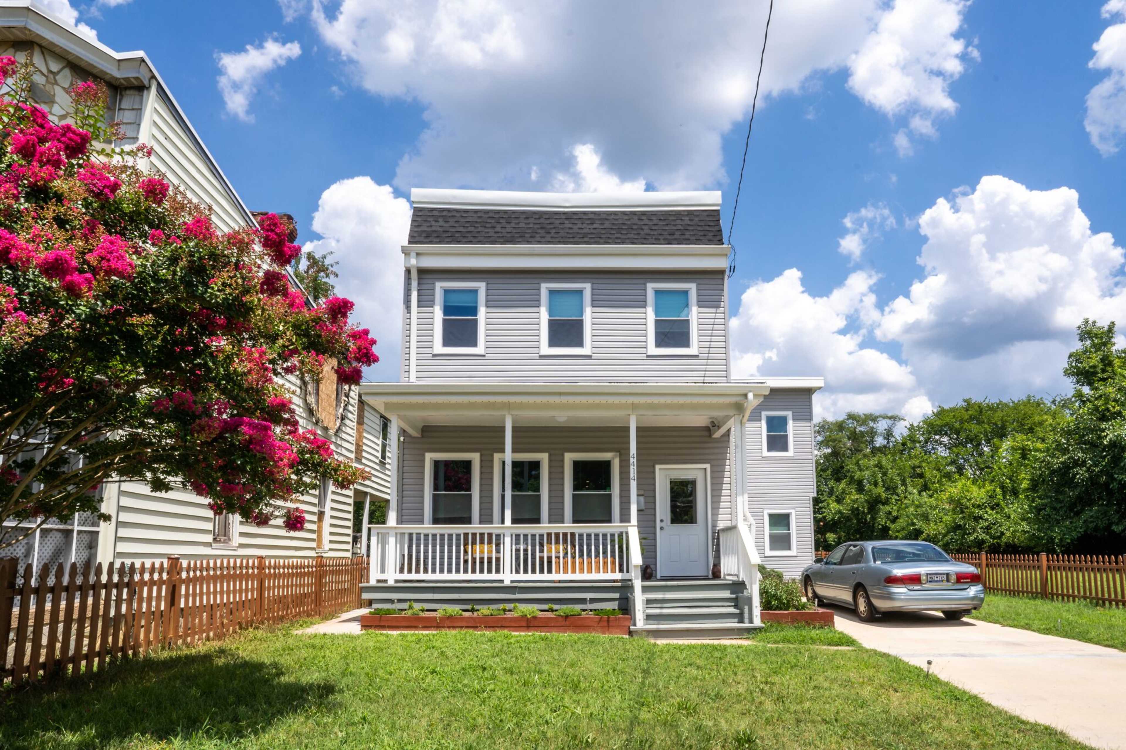 A two-story house with a porch and flowered shrubbery sits beside a paved driveway, under a cloudy sky.