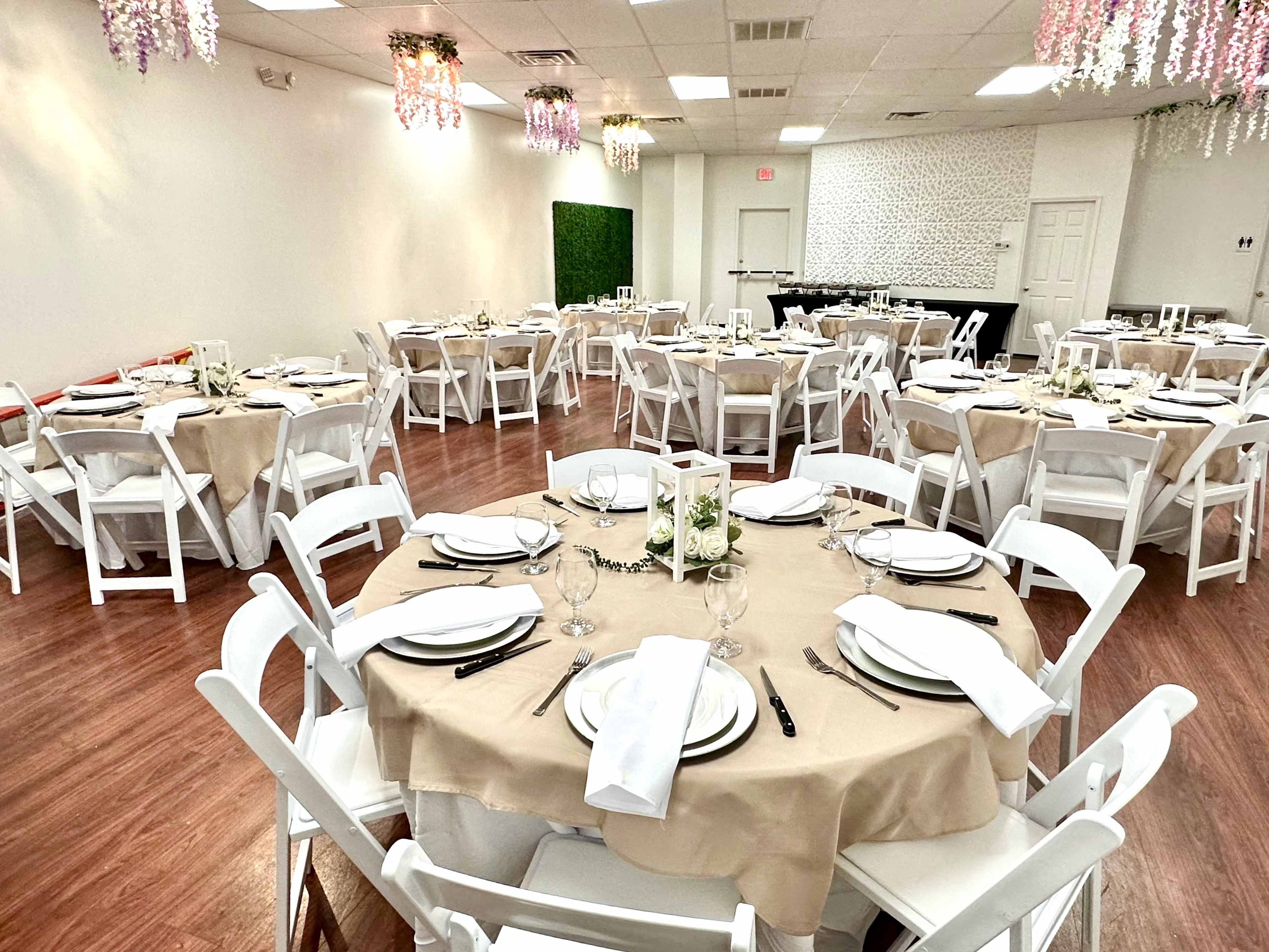 A spacious event room set with several round tables, each covered in beige tablecloths and arranged with white dinnerware and glassware, surrounded by white chairs.