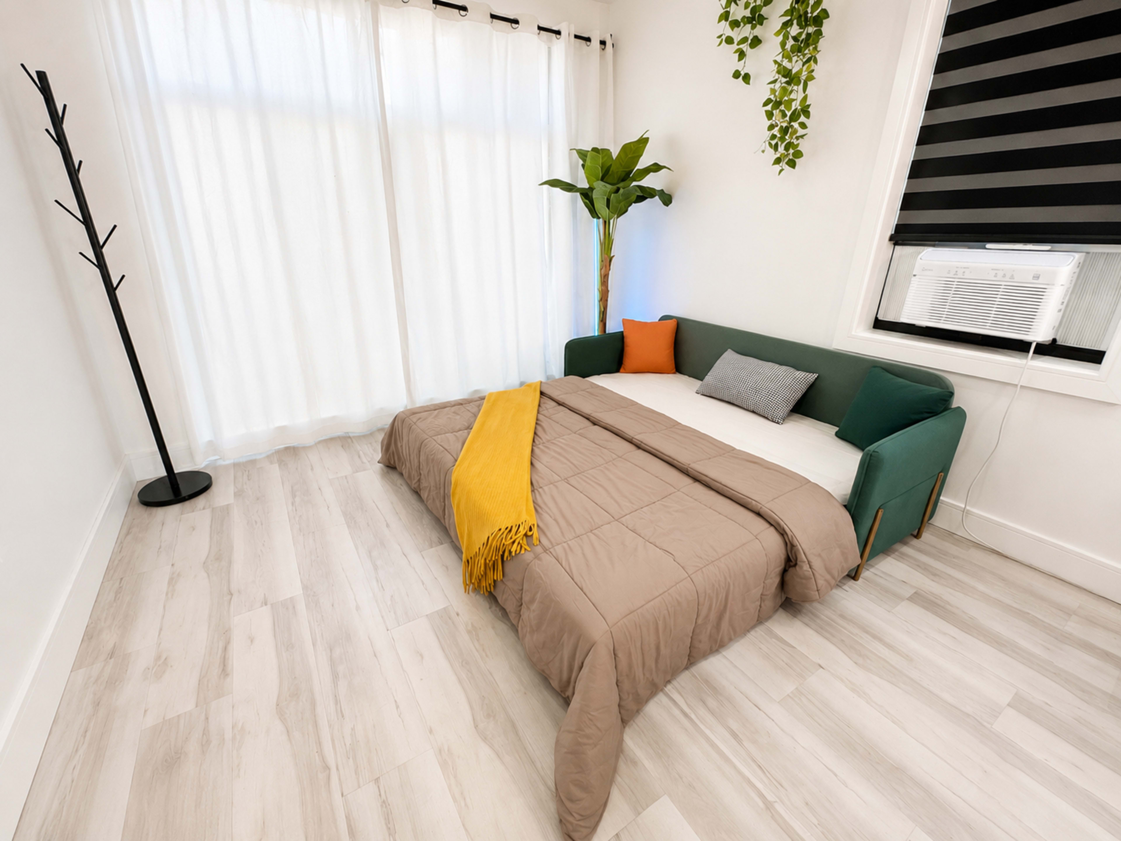 A cozy room features a bed on the floor with a green sofa and a standing coat rack, accompanied by curtains and an air conditioning unit.