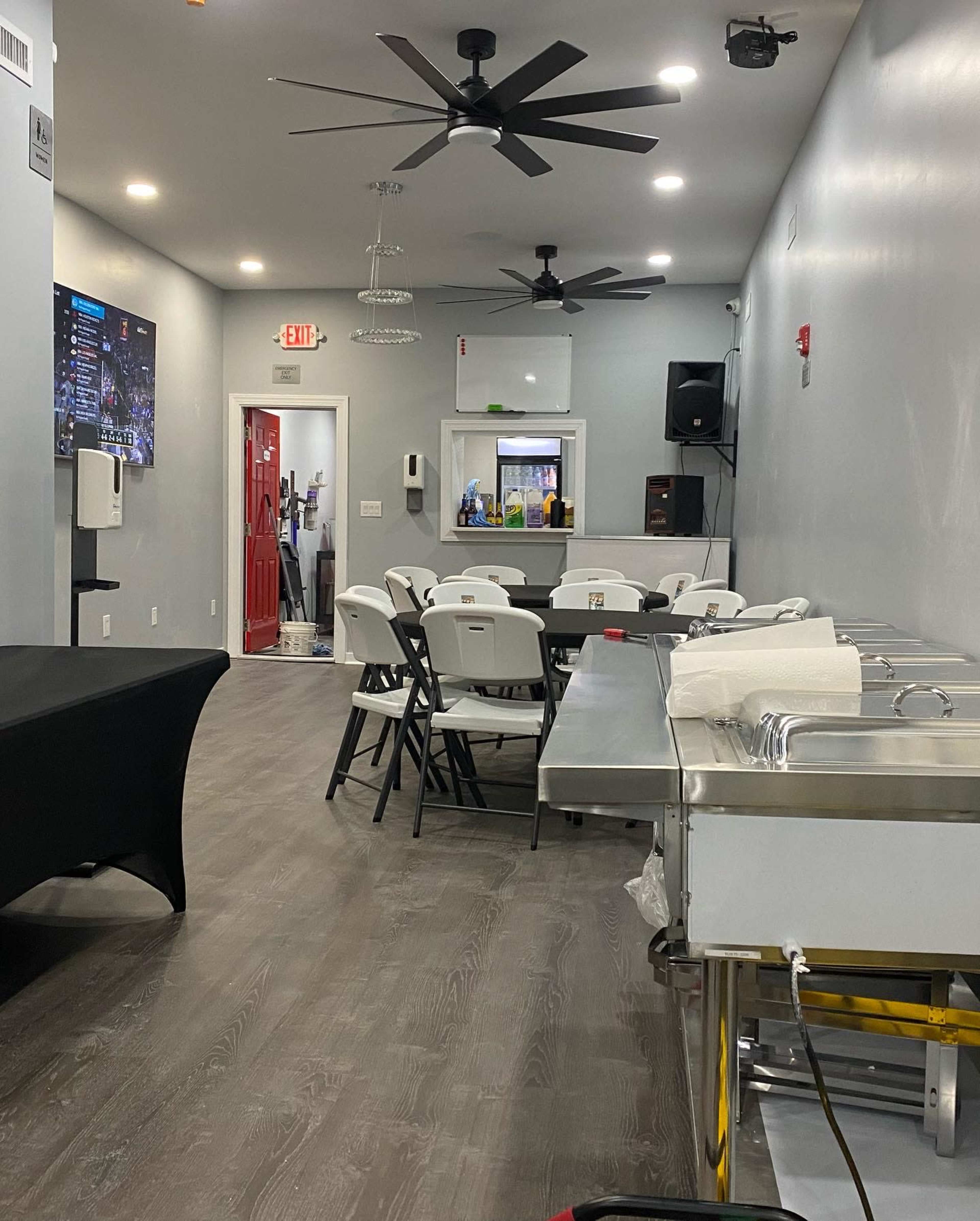 The image shows a spacious indoor area with a row of white tables and chairs, a serving counter, and a red door at the back.