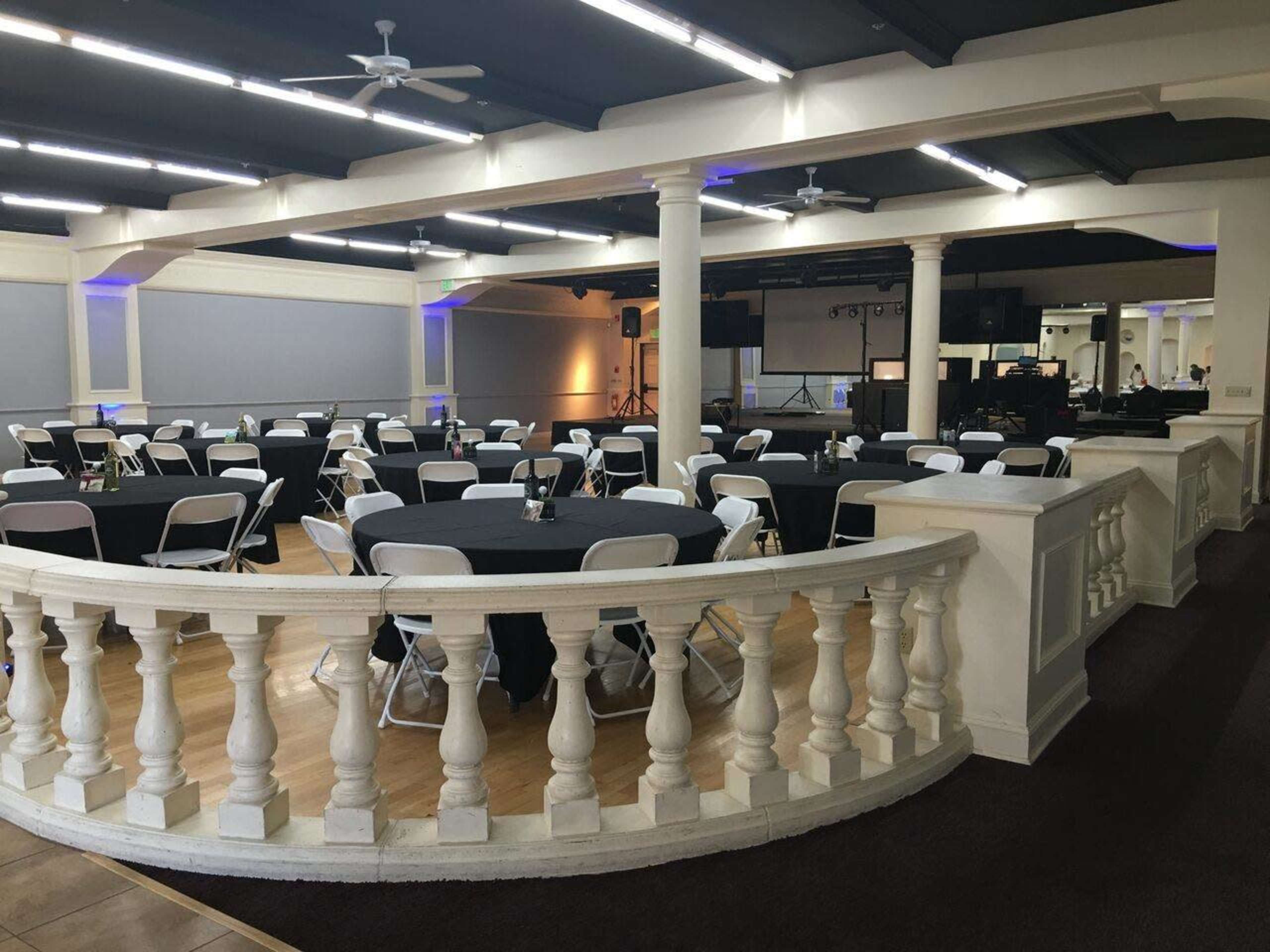 The image shows a large event space with round tables covered in black tablecloths, white folding chairs, and columns along the perimeter.