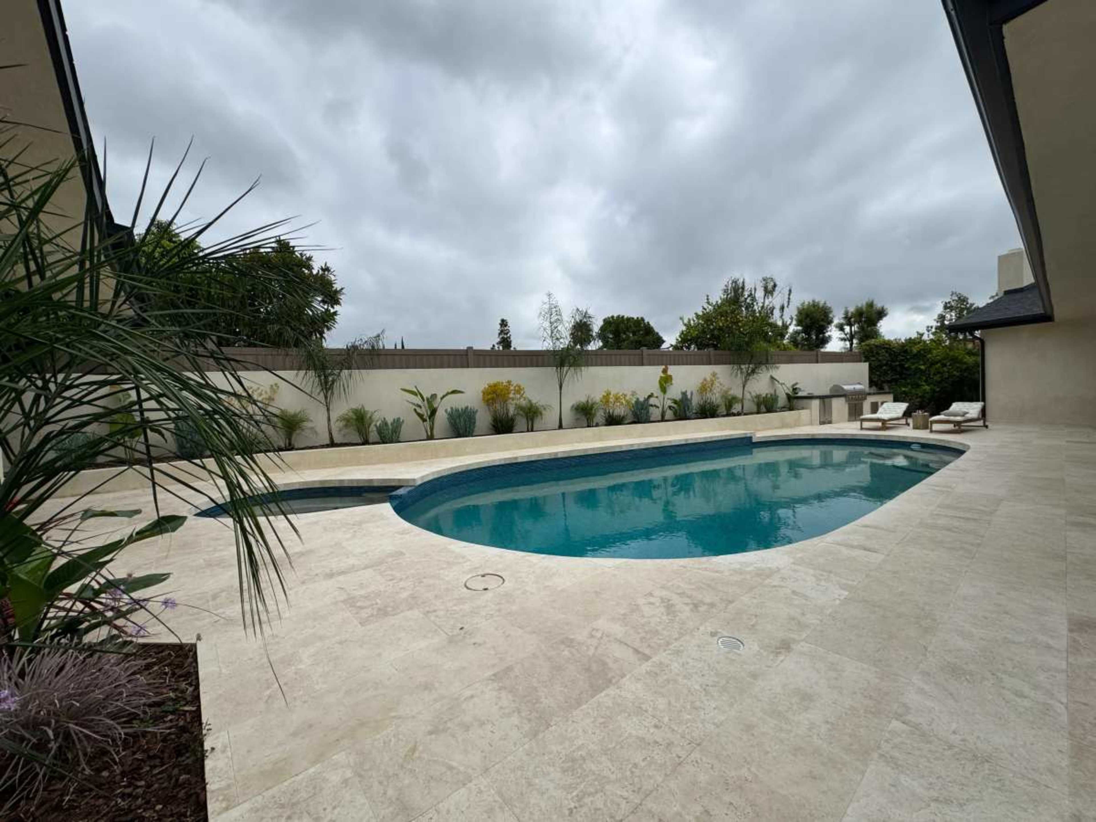 A rectangular swimming pool is surrounded by a stone patio and landscaped greenery under a cloudy sky.