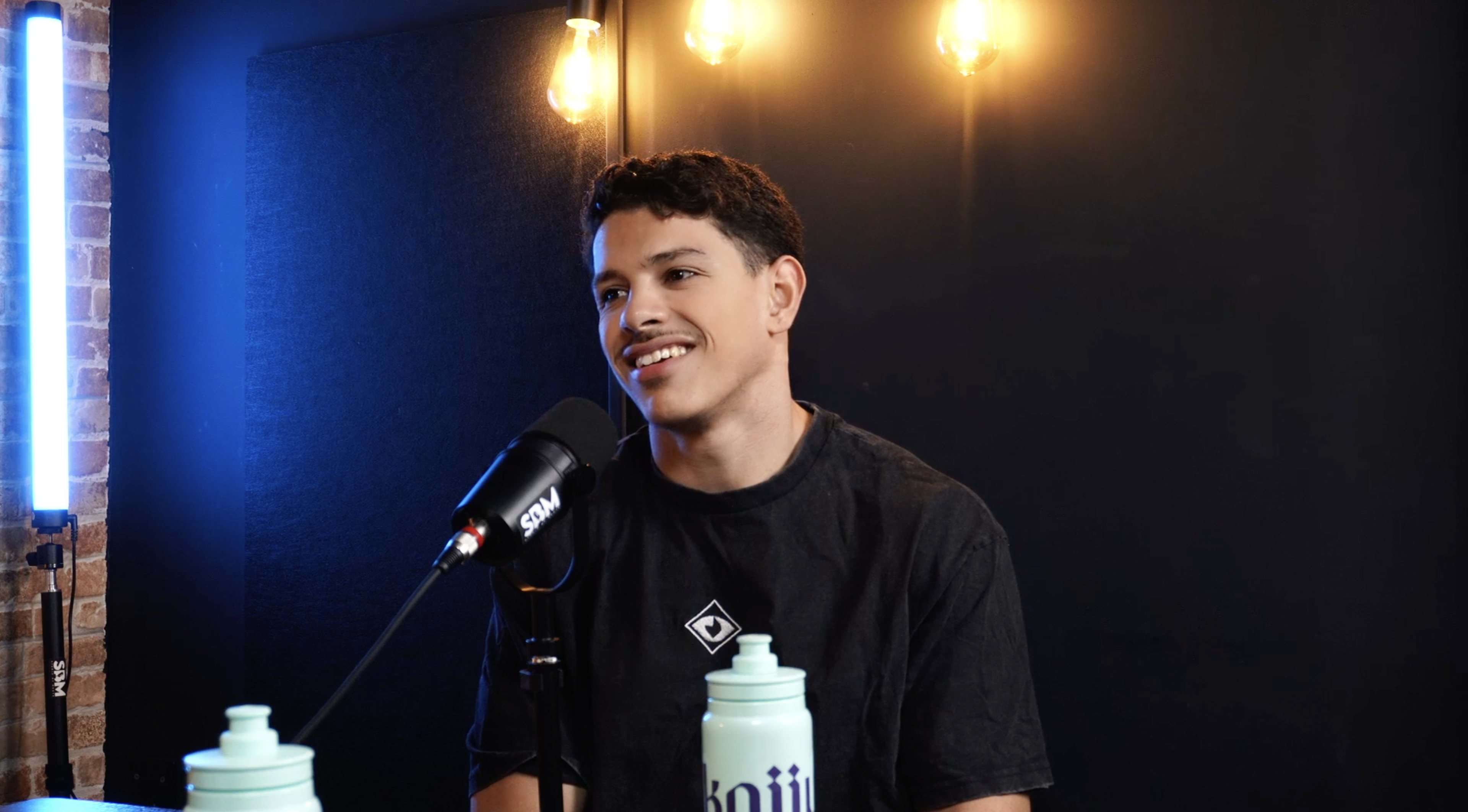 A young man with curly hair is sitting at a microphone, smiling, in a studio with warm lighting and a dark backdrop.