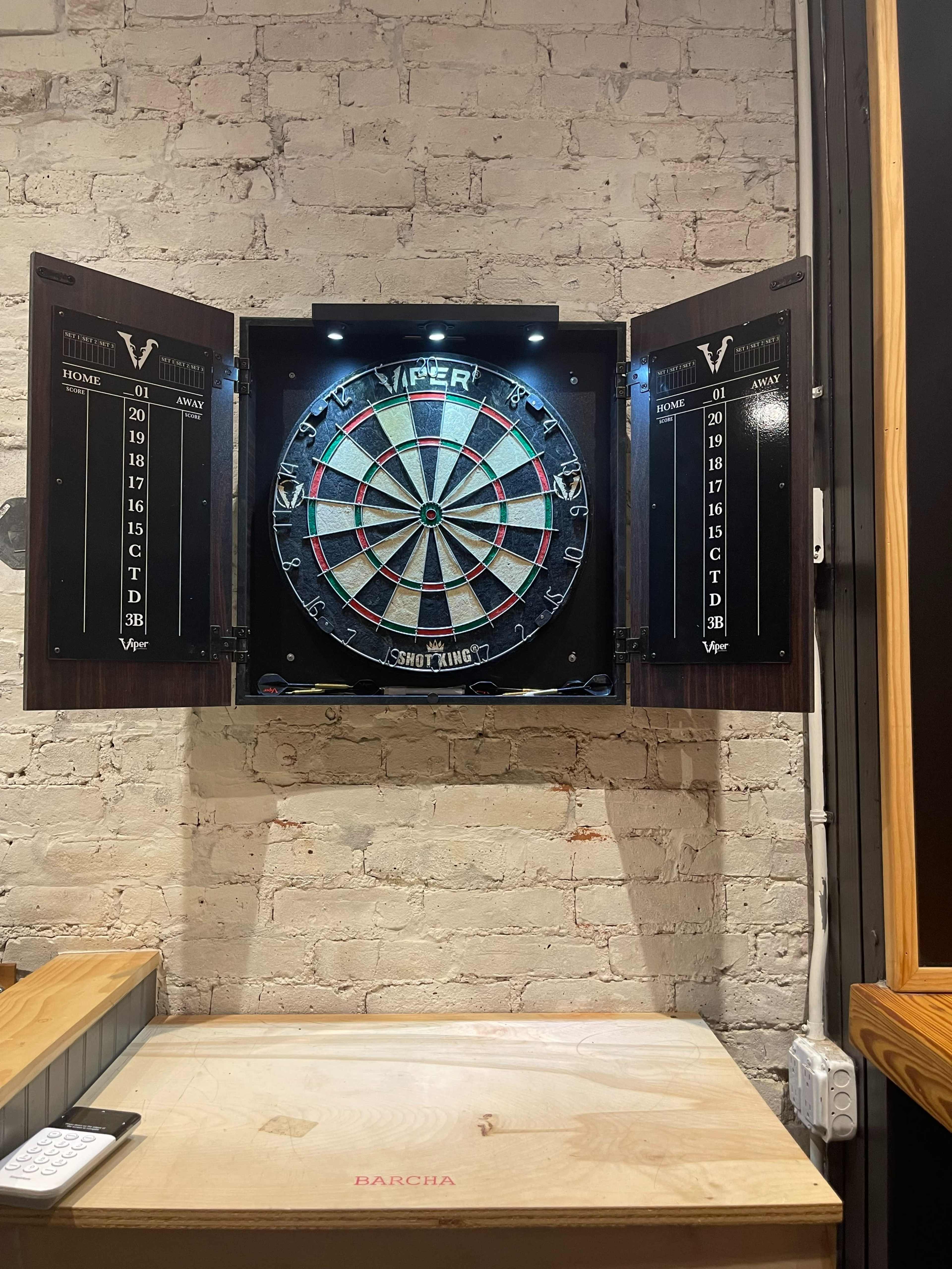 A dartboard with a wooden cabinet is mounted on a brick wall, illuminated by two small lights.