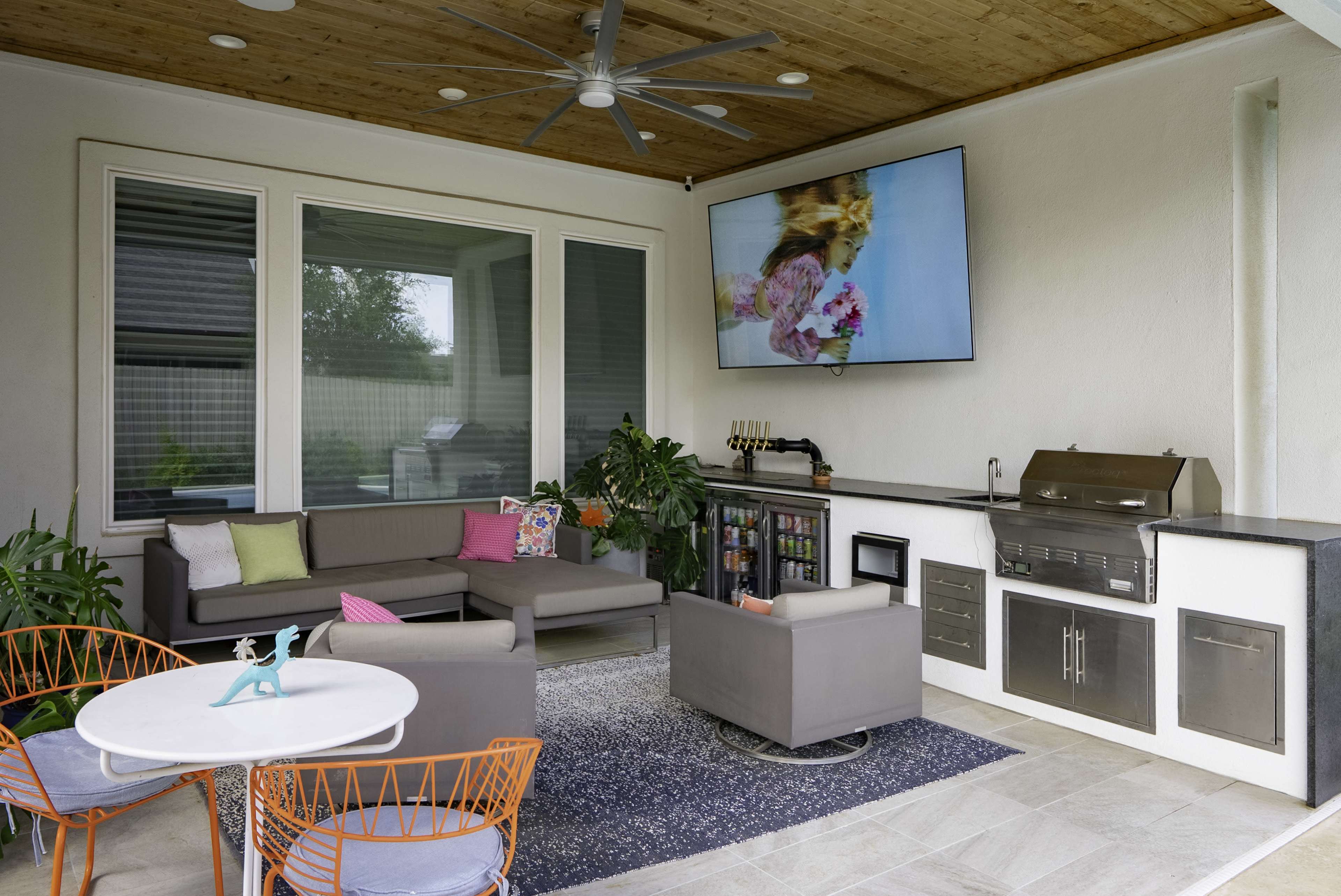 The image shows a covered outdoor patio with a sectional couch, a coffee table, a grill station, and a large screen mounted on the wall.
