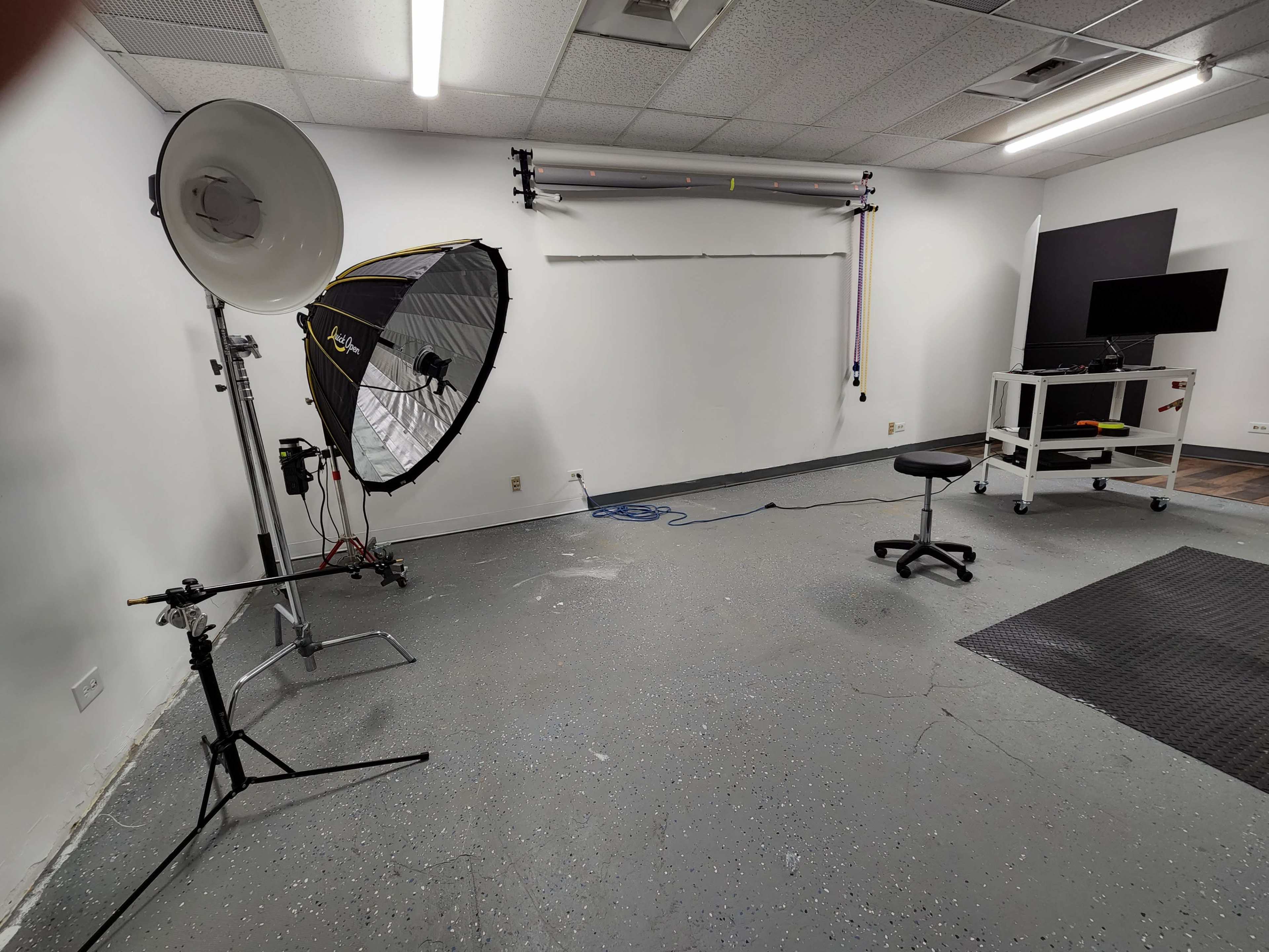 An empty photography studio featuring a large softbox light, a stool, a backdrop support system, and a mobile workstation.