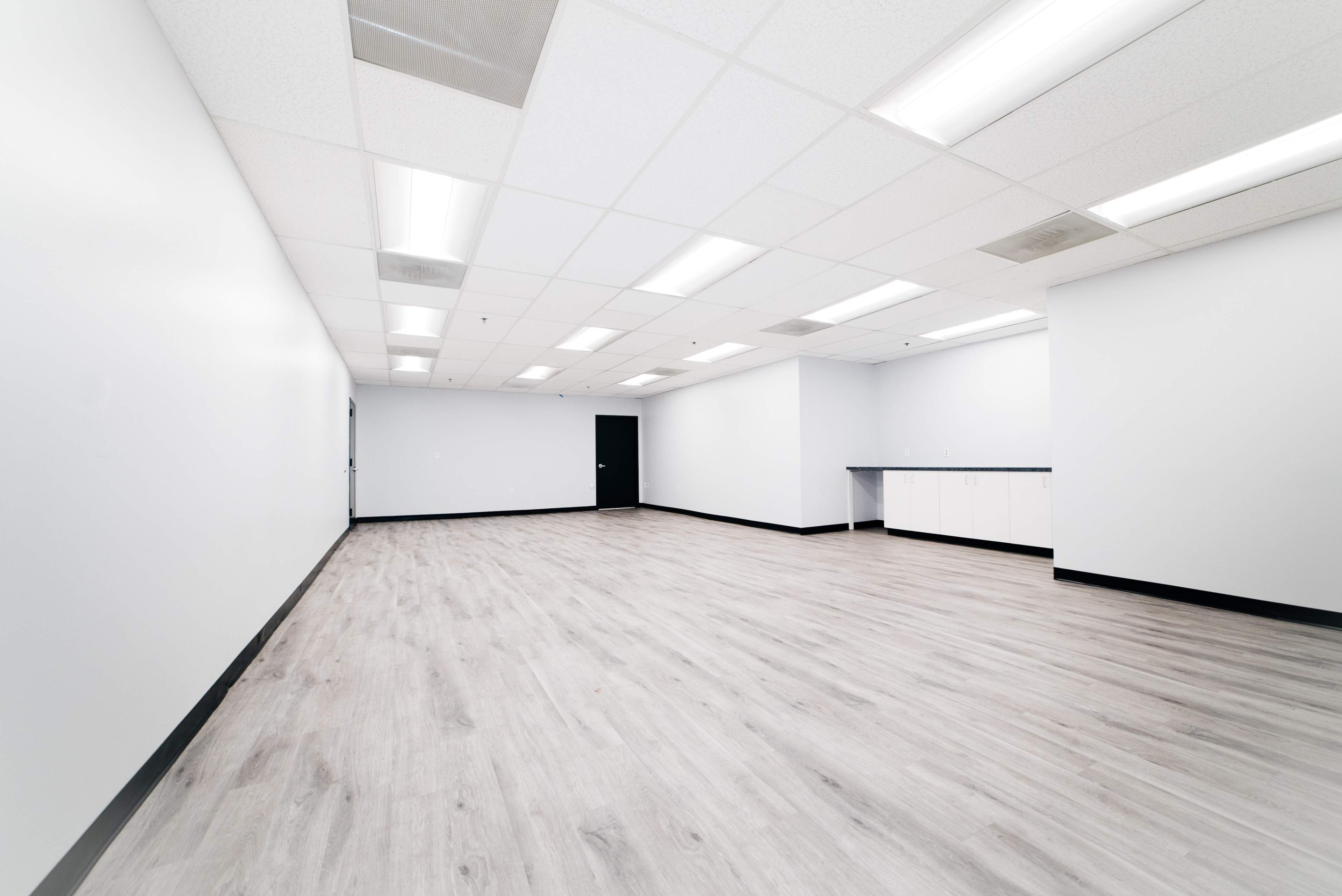 The image shows a large, empty room with gray vinyl flooring, white walls, and fluorescent lighting.