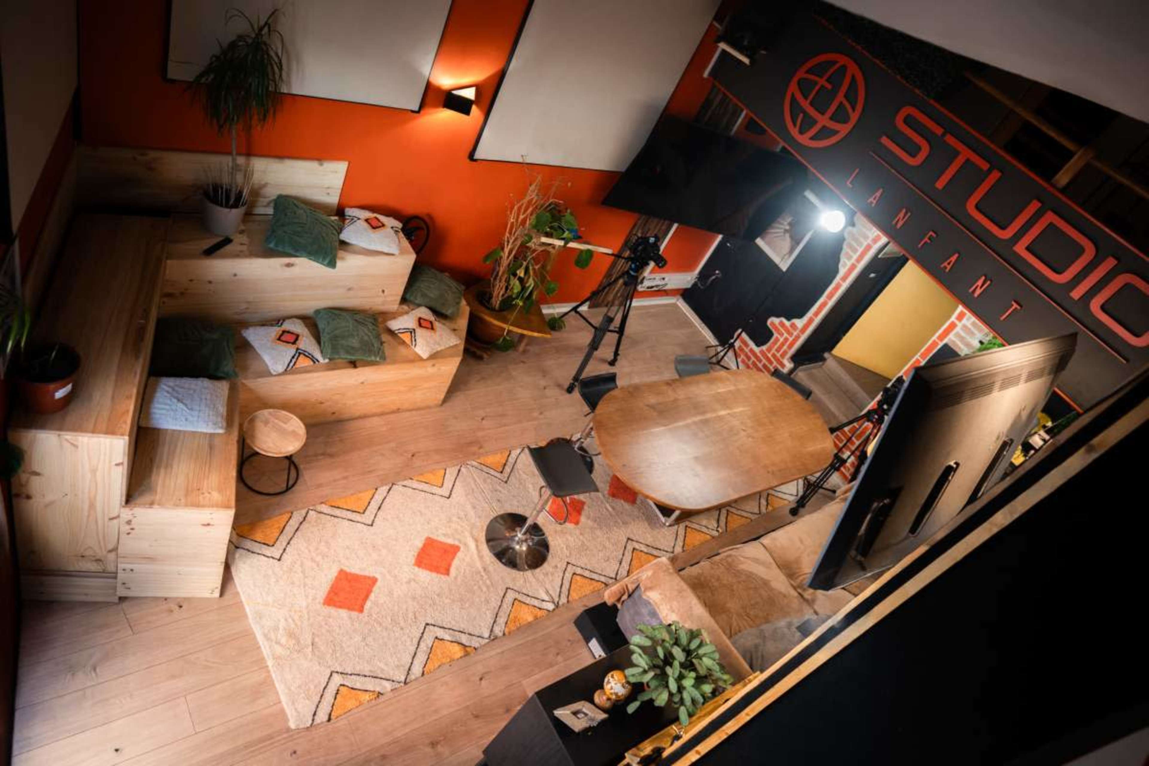 The image shows an overhead view of a modern studio space featuring wooden seating, a large table, various plants, and colorful decor elements.