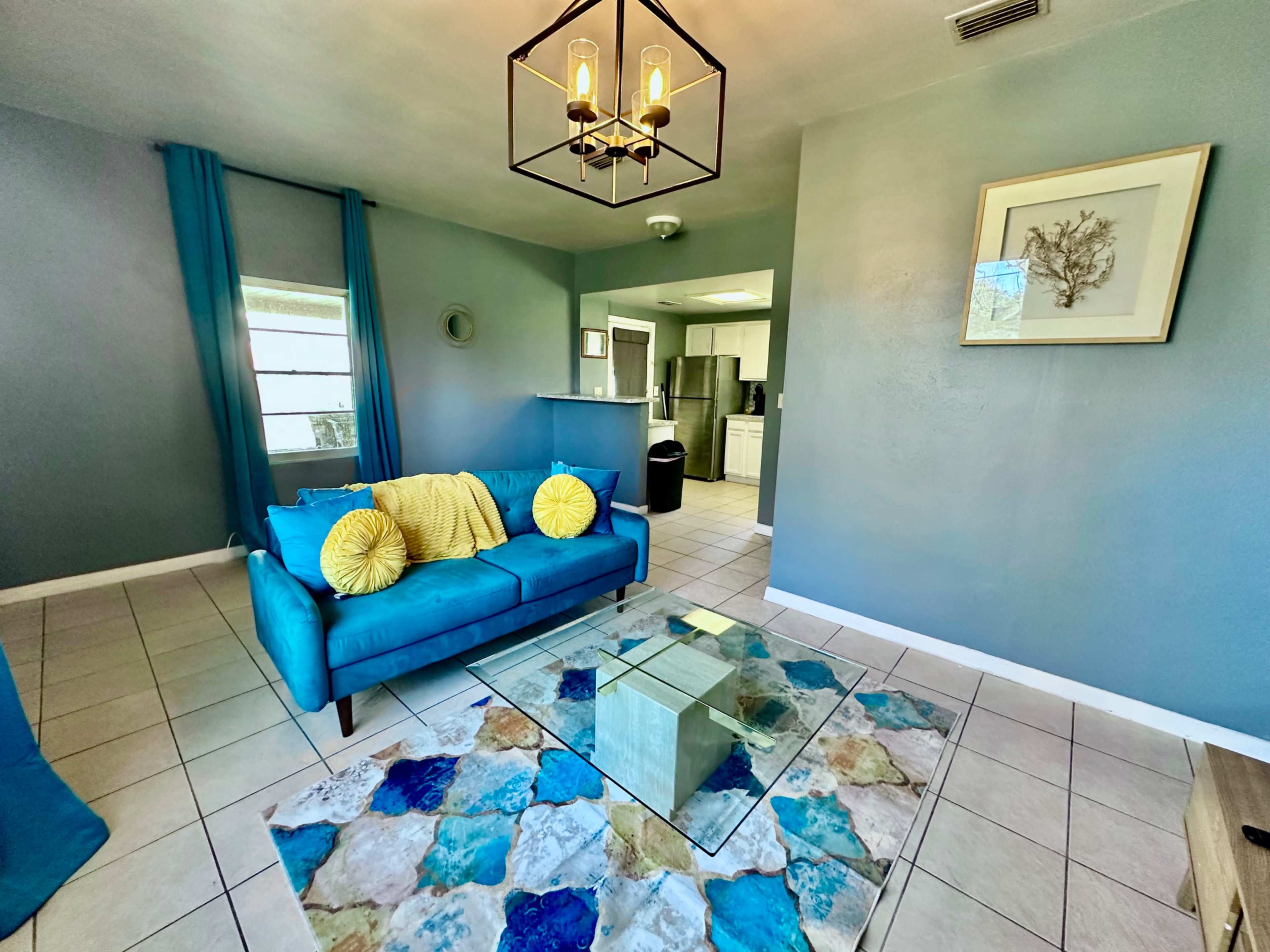 The image shows a living room with a blue sofa, yellow pillows, a glass coffee table, and light blue walls, leading to a kitchen area in the background.