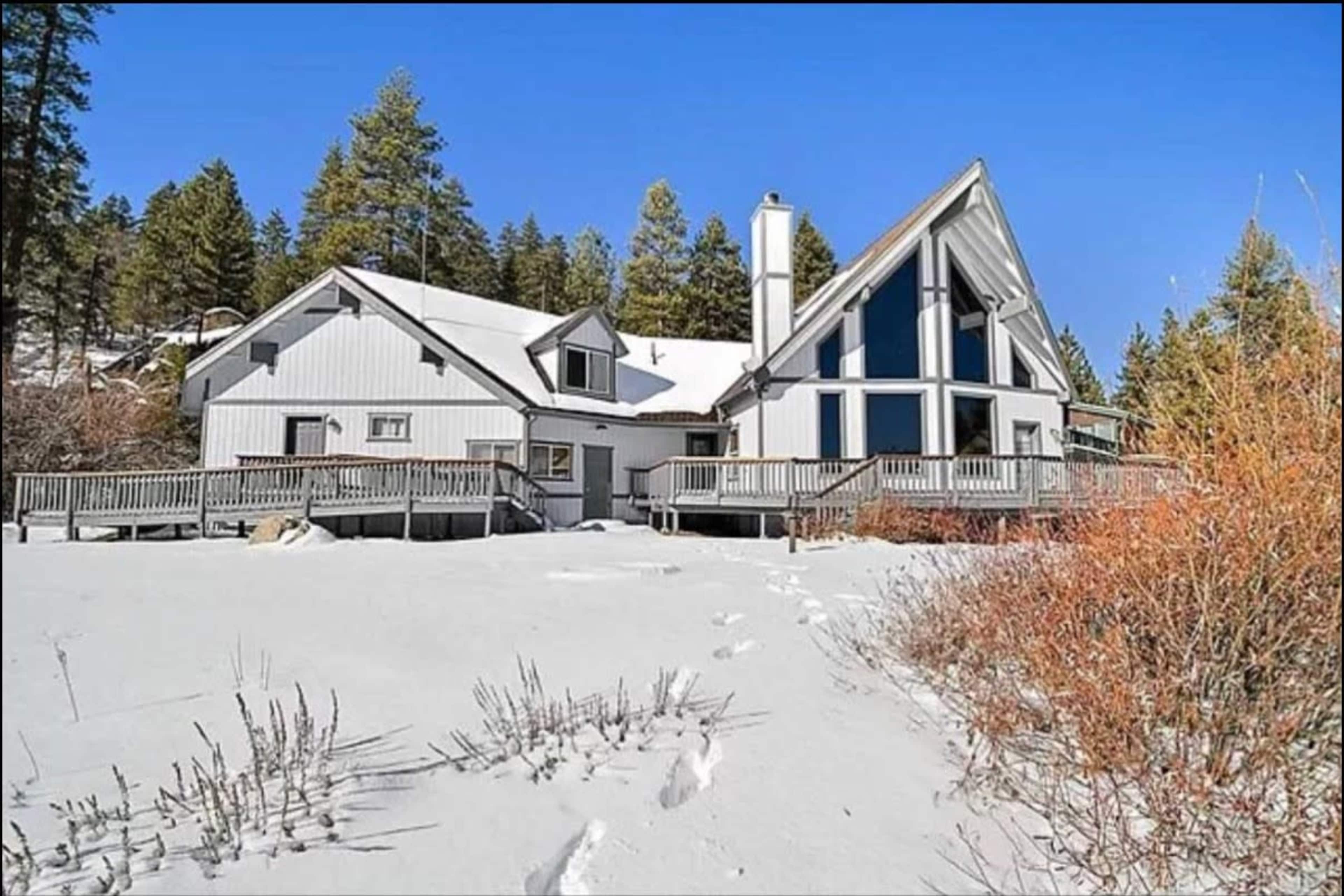 A large, modern house with a distinctive A-frame roof sits in a snowy landscape surrounded by evergreen trees.