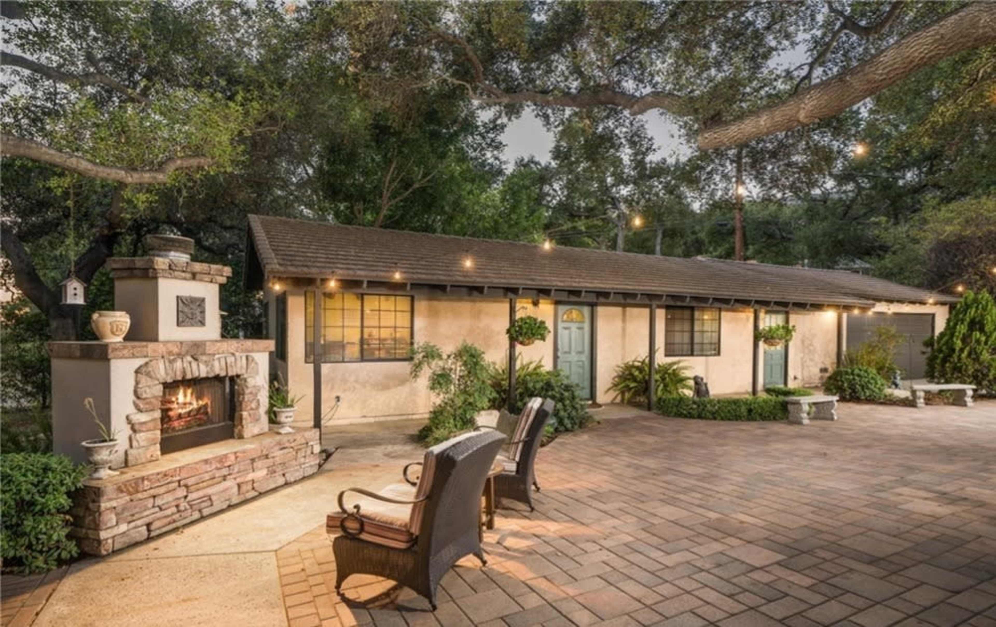 The image shows a cozy exterior of a cottage-style home with a stone fireplace and patio seating under string lights, surrounded by greenery.
