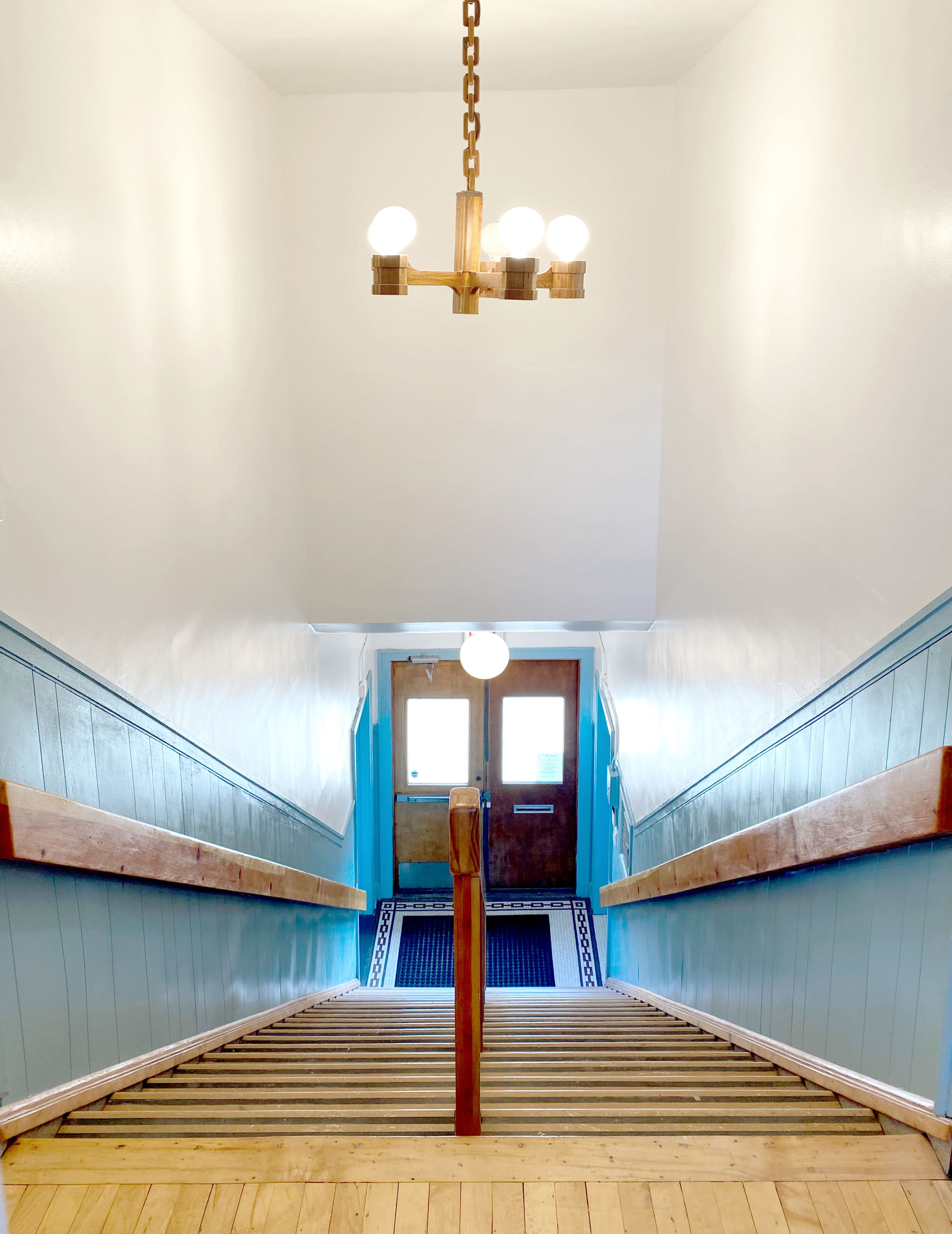 The image shows a well-lit stairwell with wooden steps leading downward to a door at the bottom.
