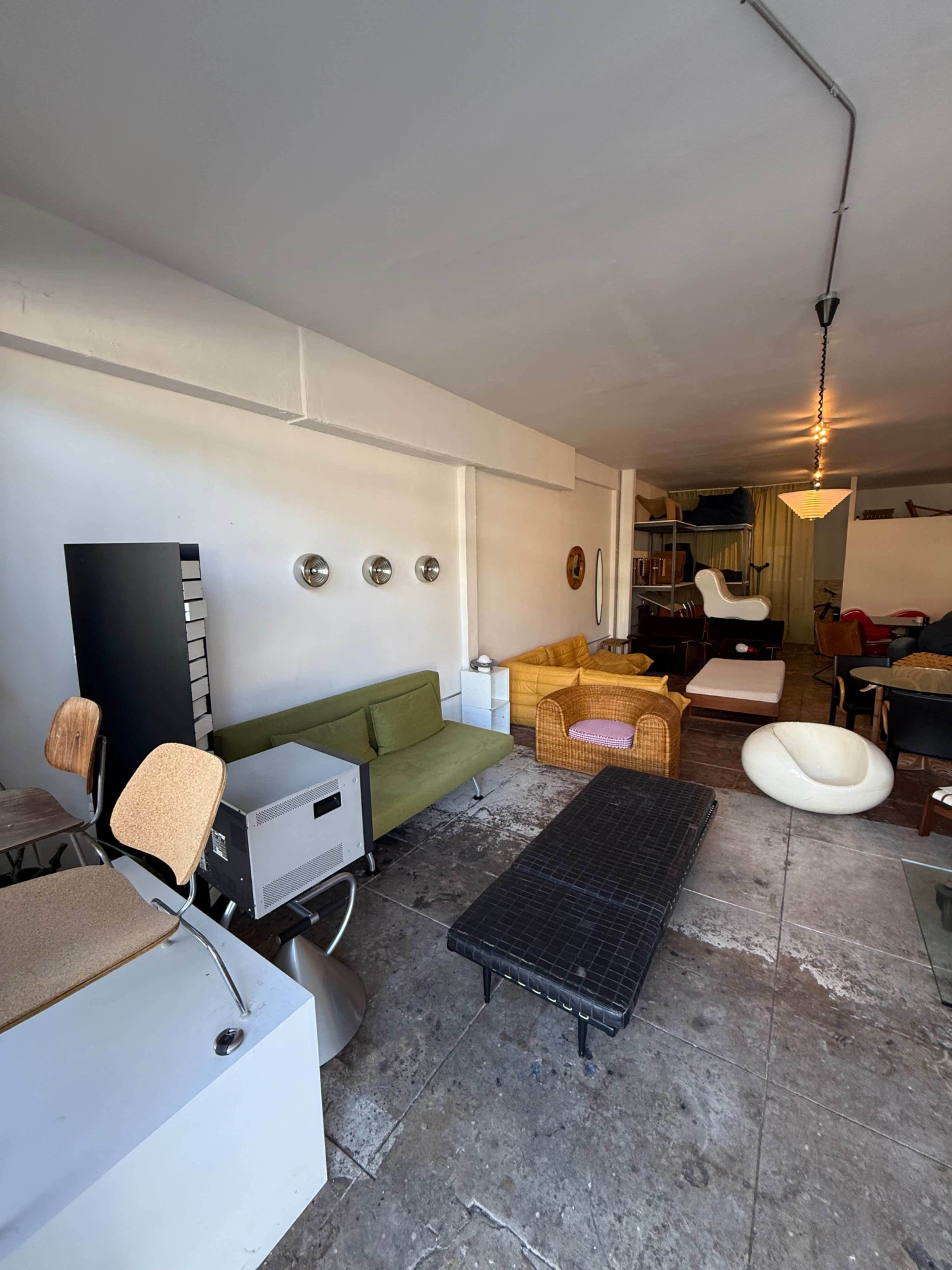The image shows a room with various pieces of furniture, including a green sofa, a wicker chair, a black coffee table, and several other seating options arranged against white walls.