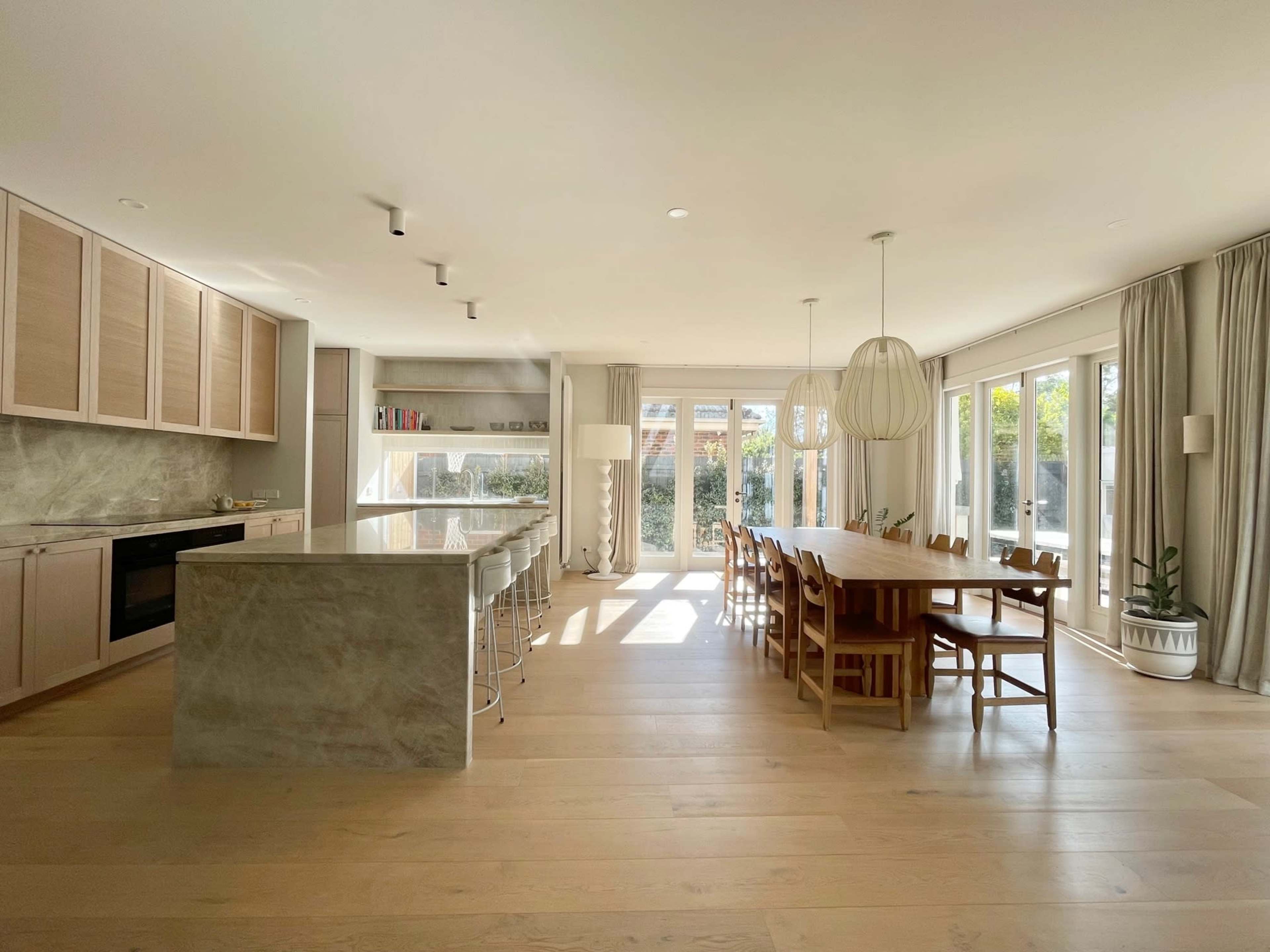 A spacious, modern kitchen and dining area with a large island, wooden dining table, and ample natural light from large windows.