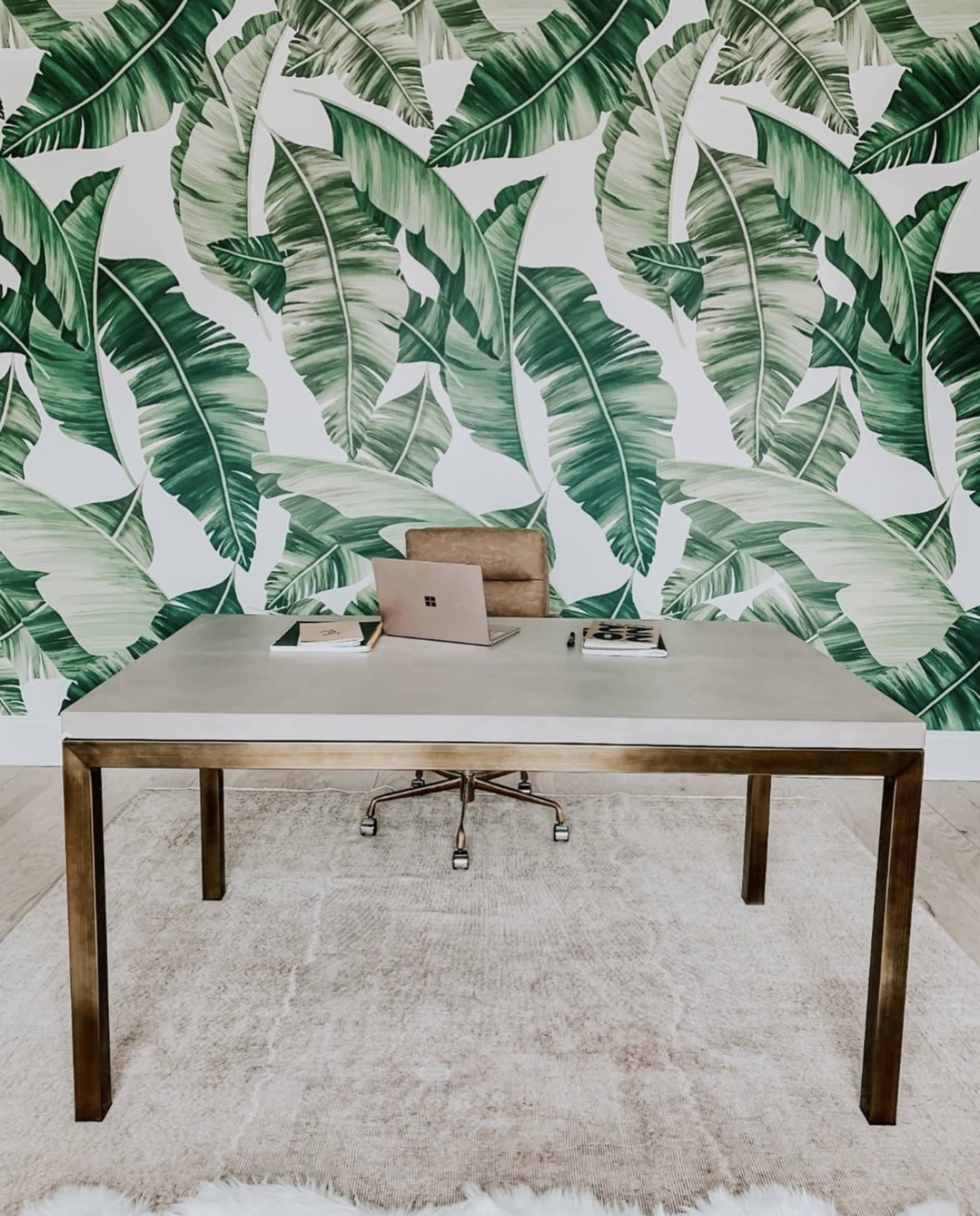 A modern workspace features a concrete desktop and a brown chair against a backdrop of large green leaf wallpaper.