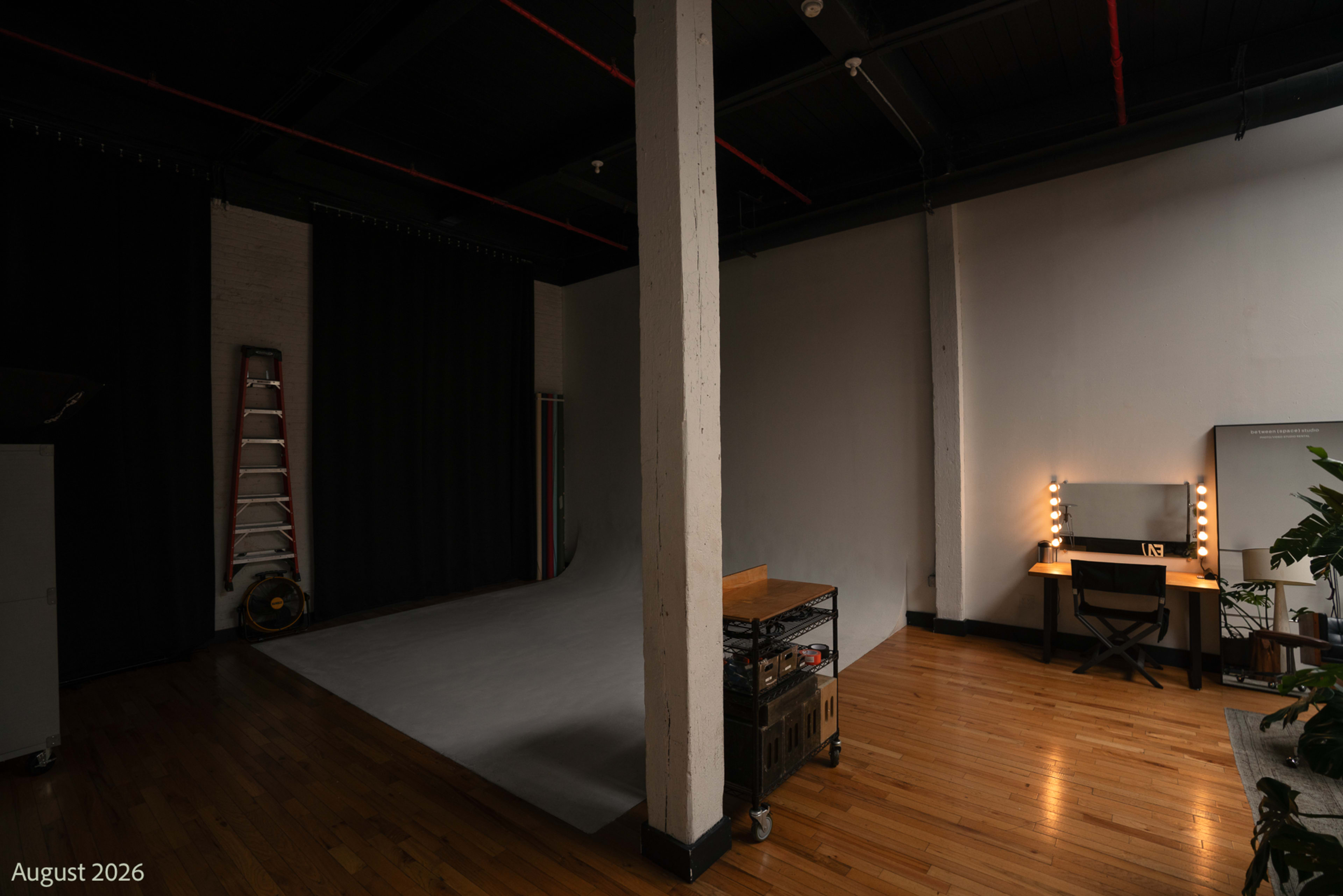 The image shows a spacious photography studio featuring a backdrop area, a makeup station with lights, and a ladder against the wall.