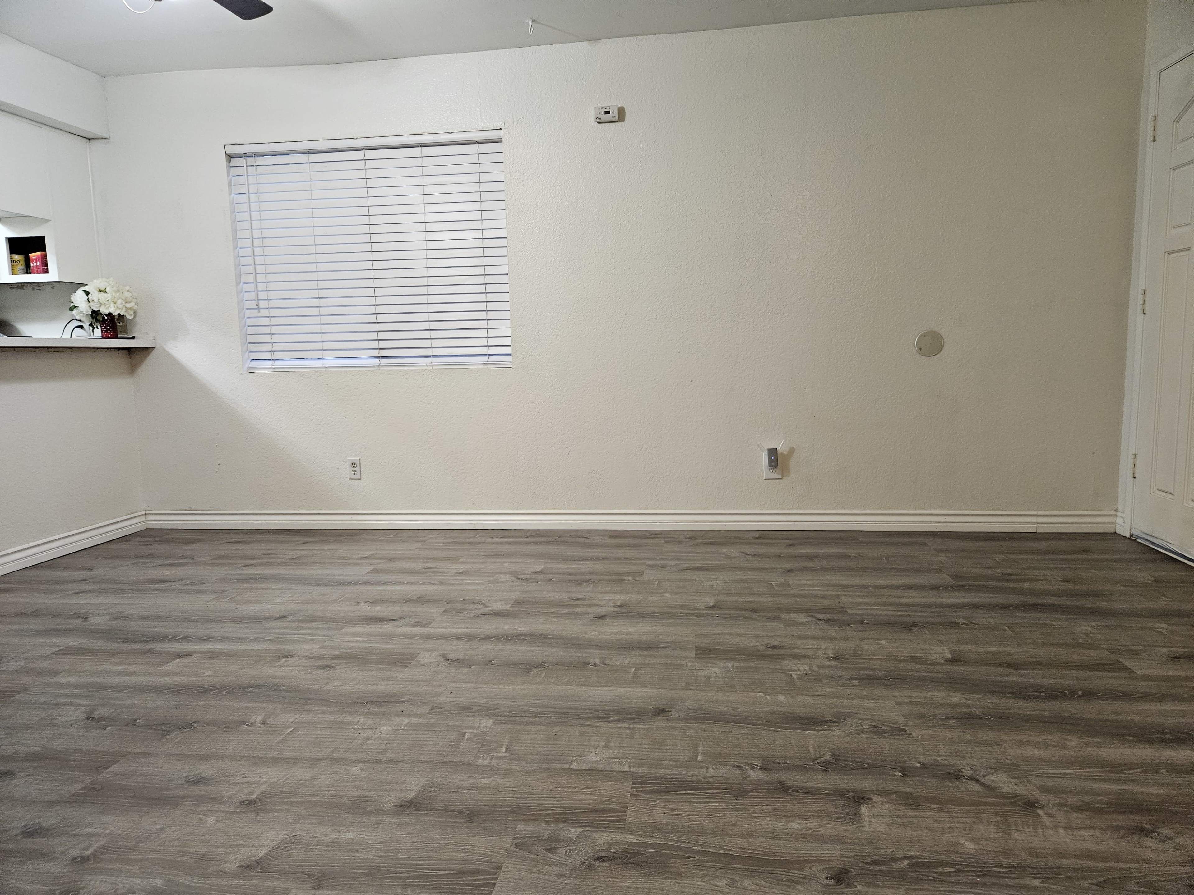 A sparsely furnished room with light-colored walls, a wooden floor, and a window covered by blinds.
