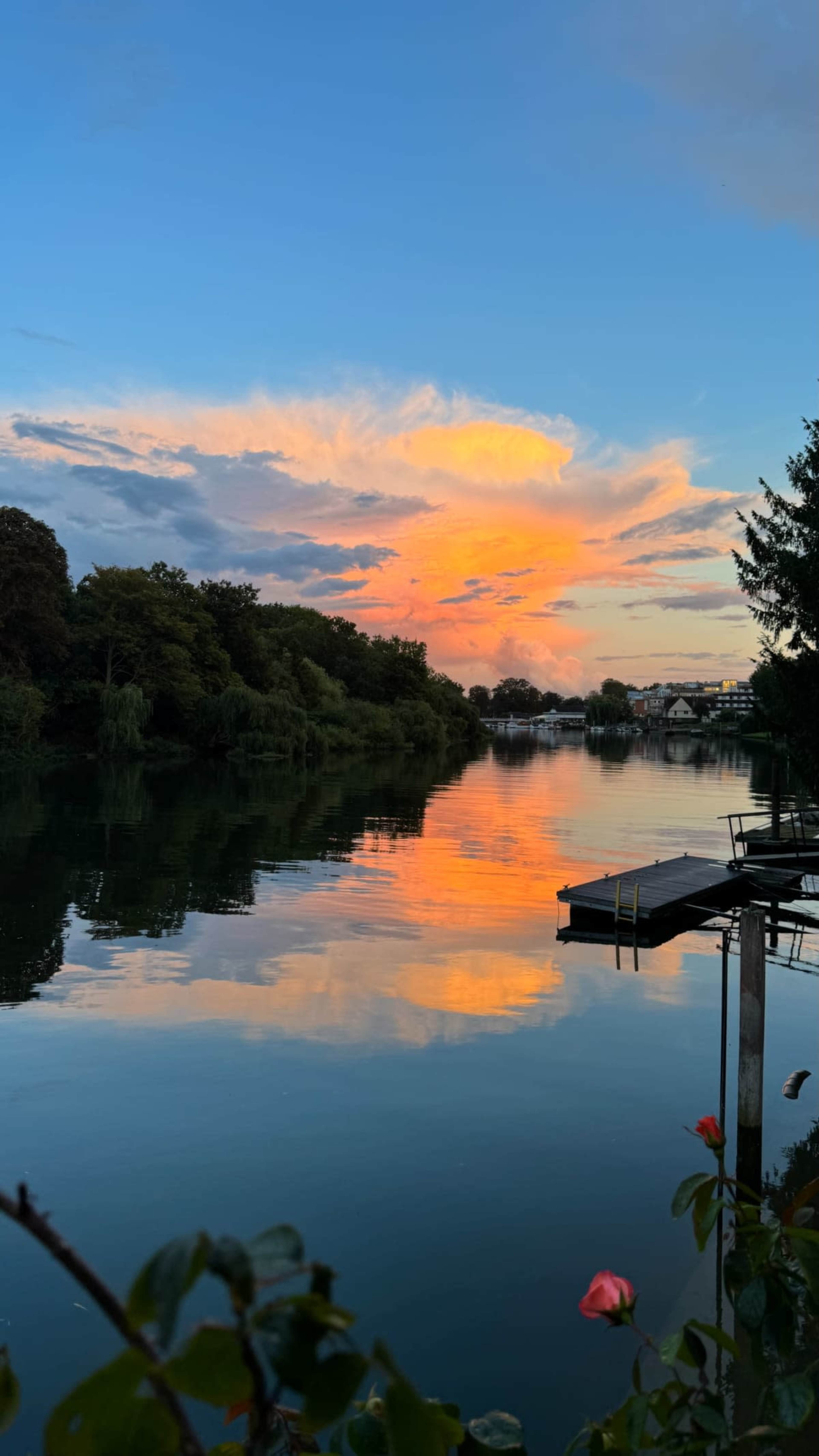 The scene shows a calm river reflecting colorful clouds and a sunset, framed by trees and a dock along the shore.