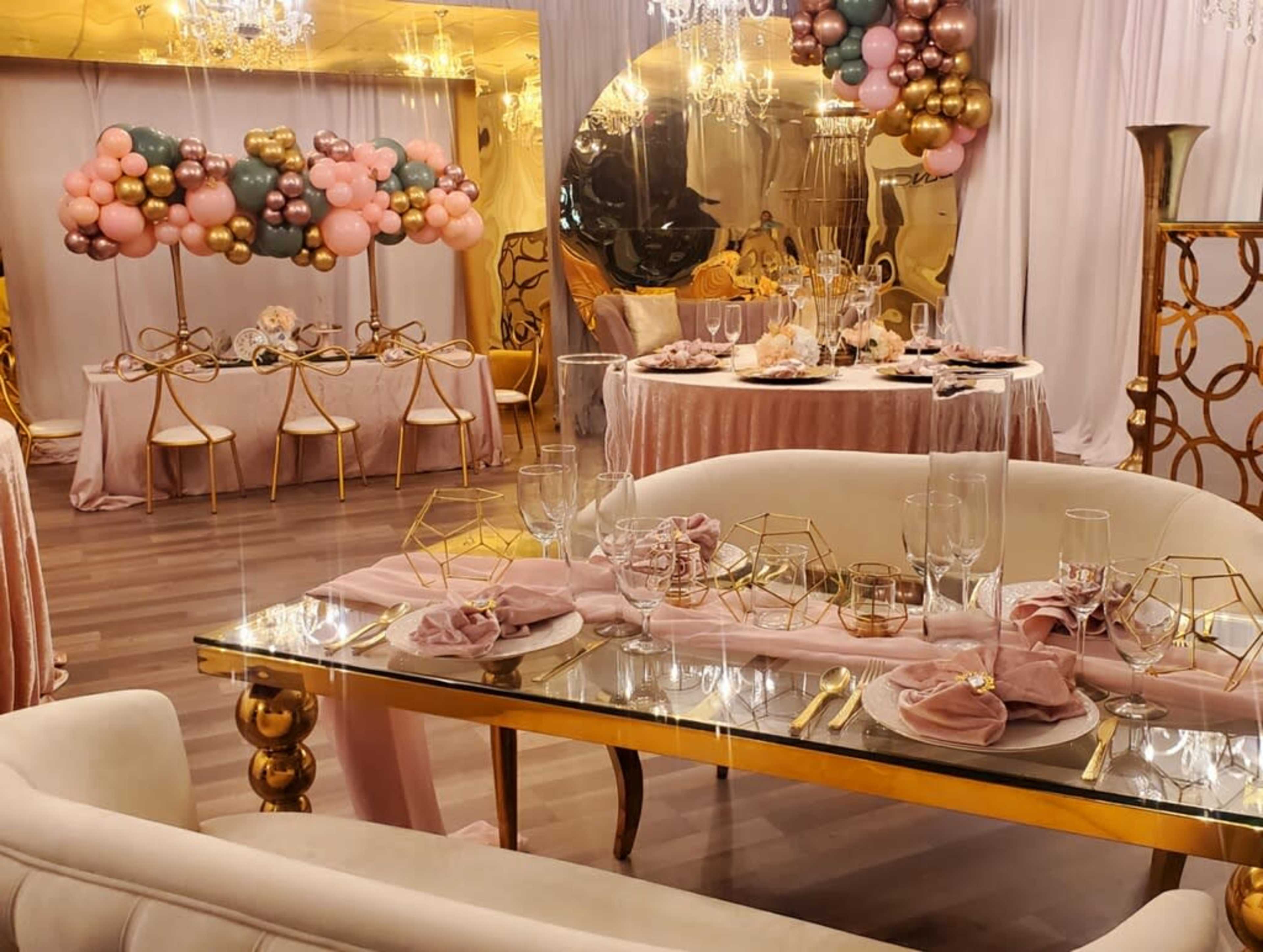 A decorated banquet hall featuring a dining table set with gold accents, pink napkins, and a backdrop of colorful balloons.