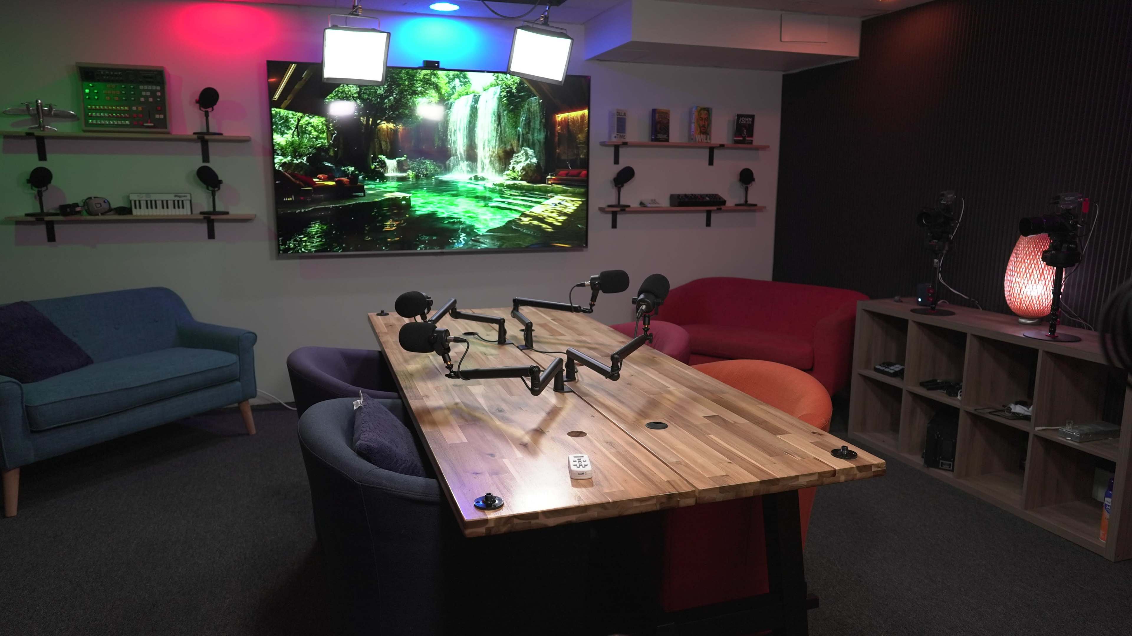 A modern room set up for recording, featuring a large screen displaying a waterfall scene, a wooden table with microphone stands, and colorful seating arrangements.