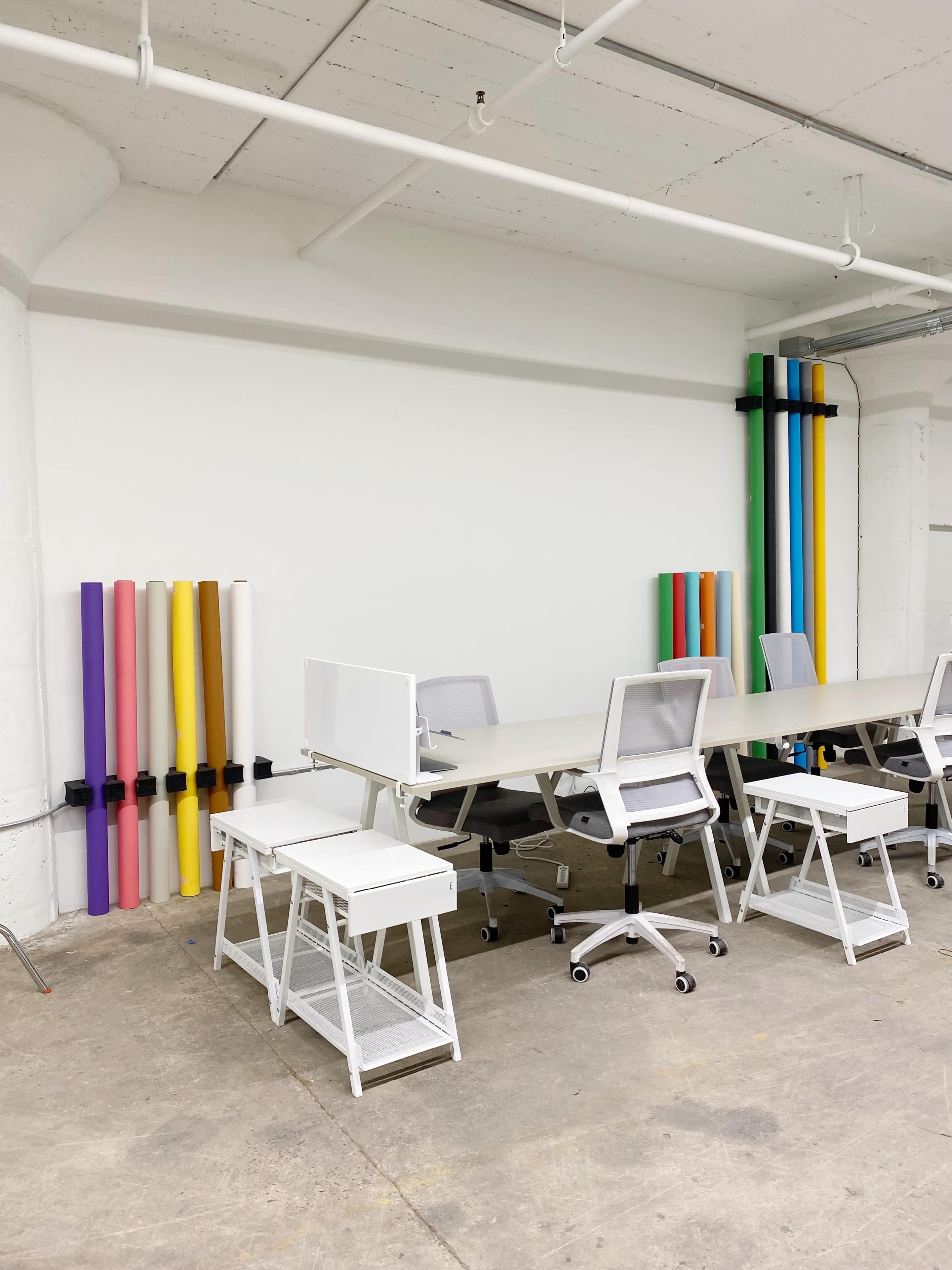 A workspace with several white desks and chairs, complemented by colorful rolls of paper stacked vertically against the wall.
