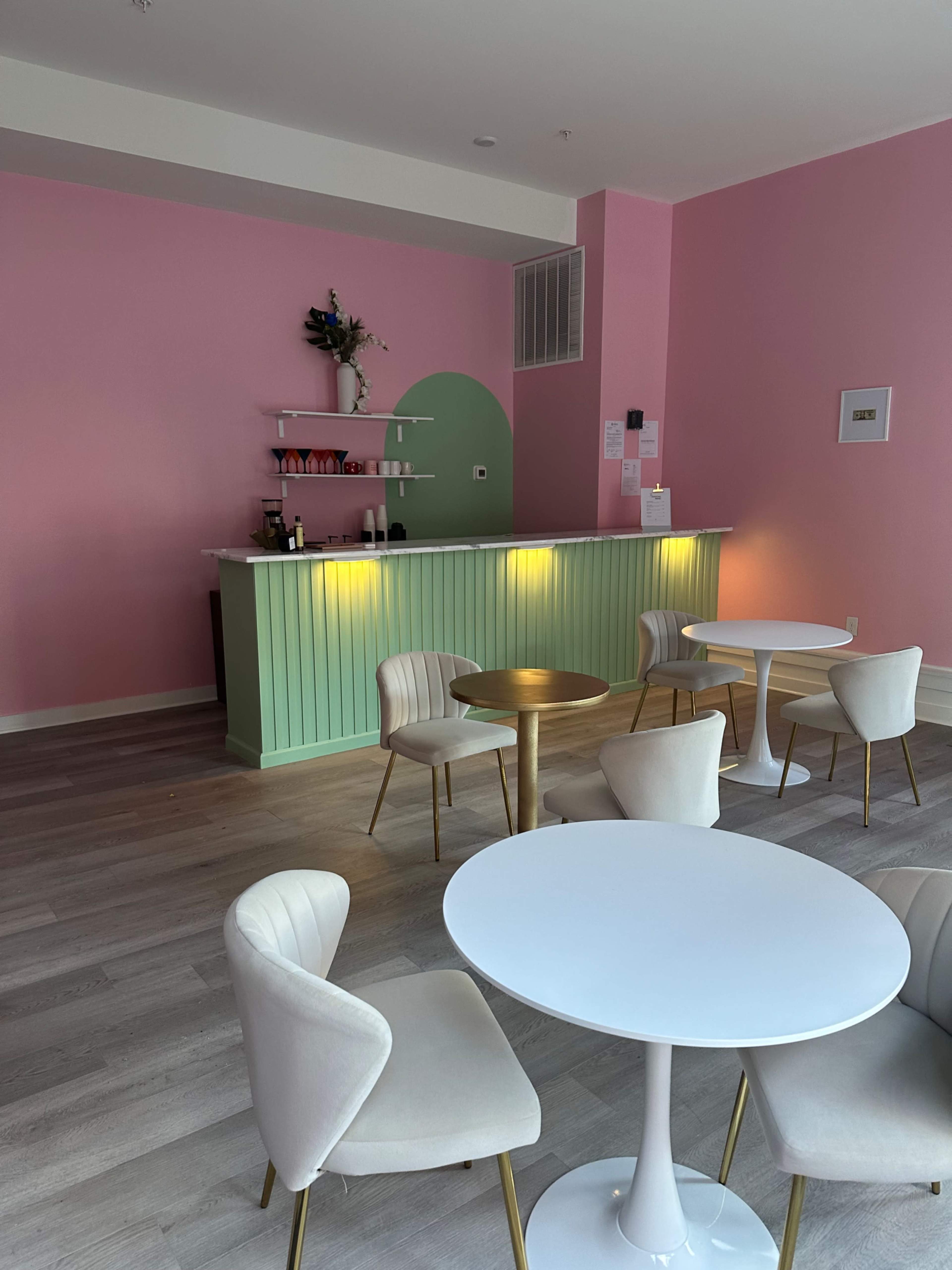 A minimalist cafe interior featuring a pink wall, a green service counter, and several round tables with white chairs.