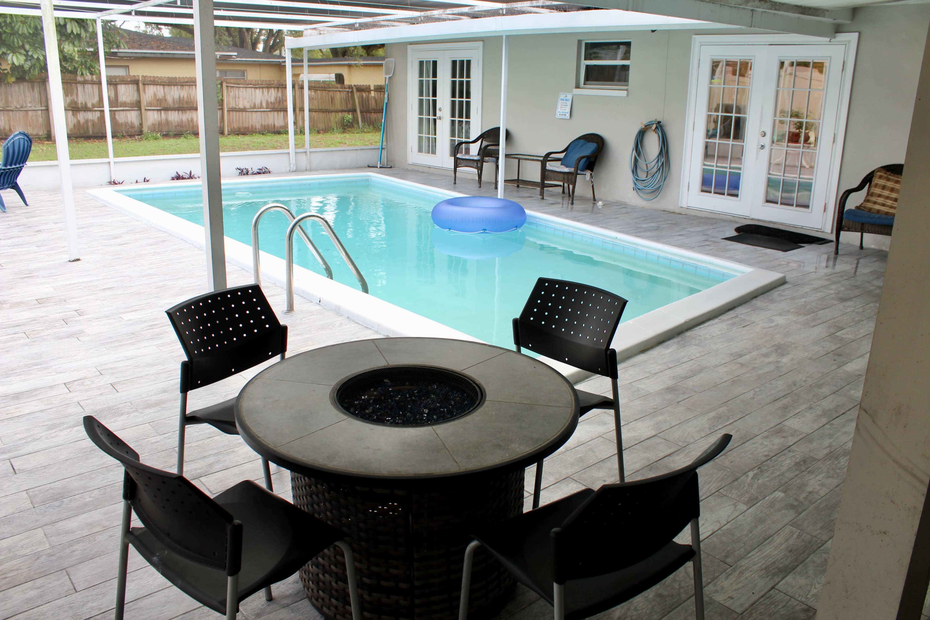 A swimming pool with a blue float, surrounded by a covered patio area featuring a circular fire pit table and black chairs.