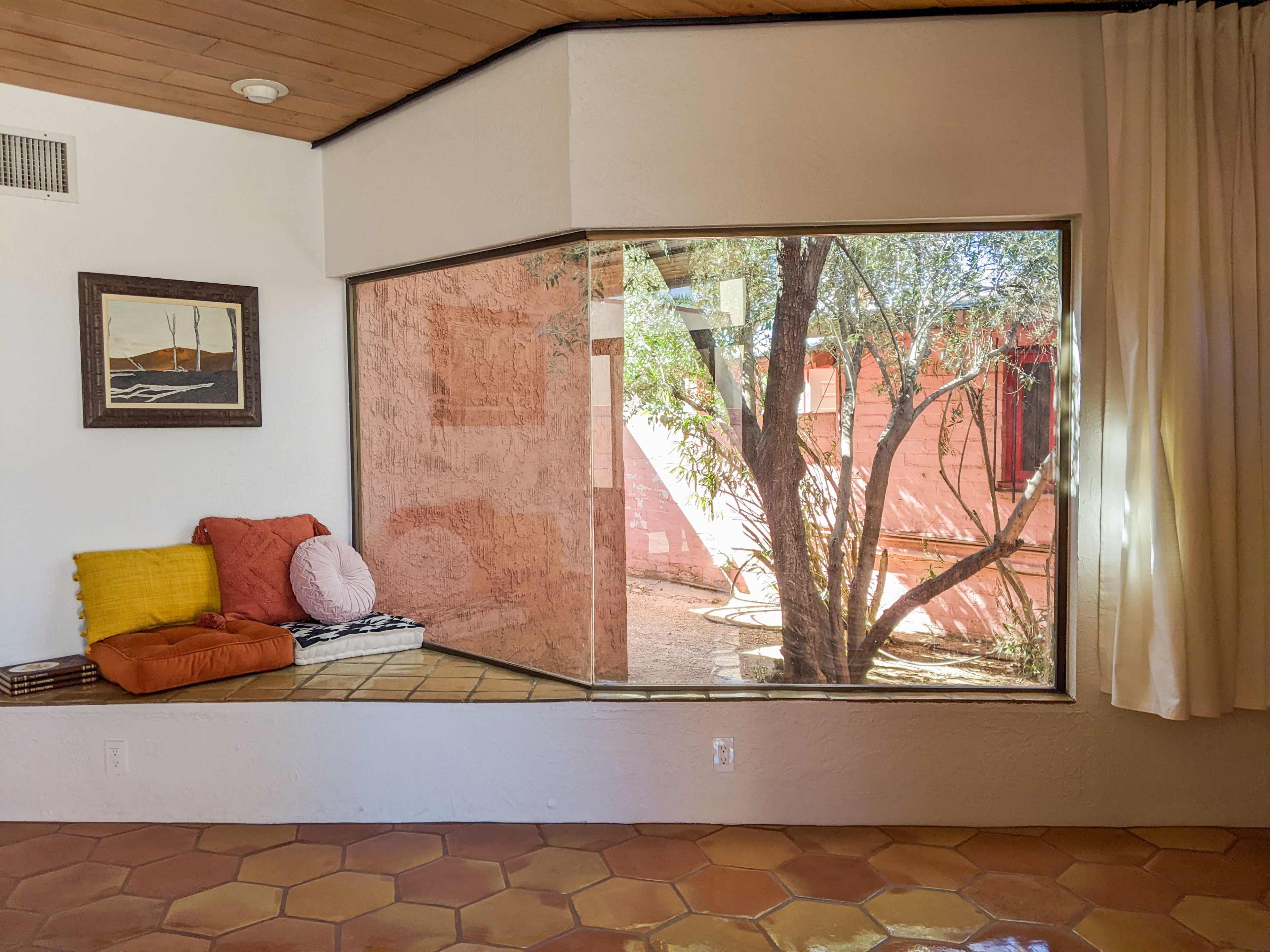A corner of a room featuring a large window with a view of an outdoor space surrounded by pink walls and greenery, along with a cushioned seating area.
