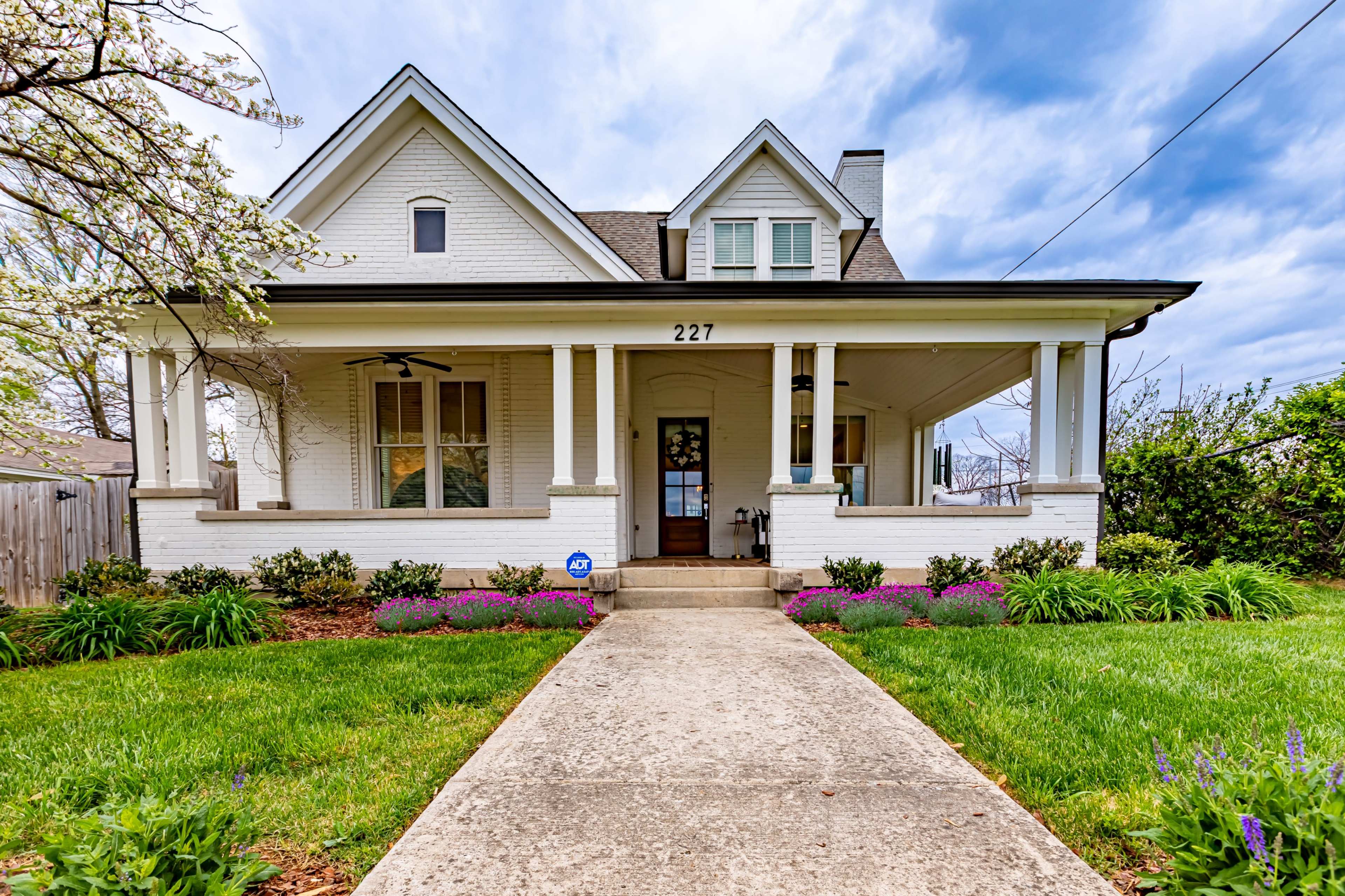 A white, two-story house with a front porch is surrounded by colorful flowers and green landscaping.
