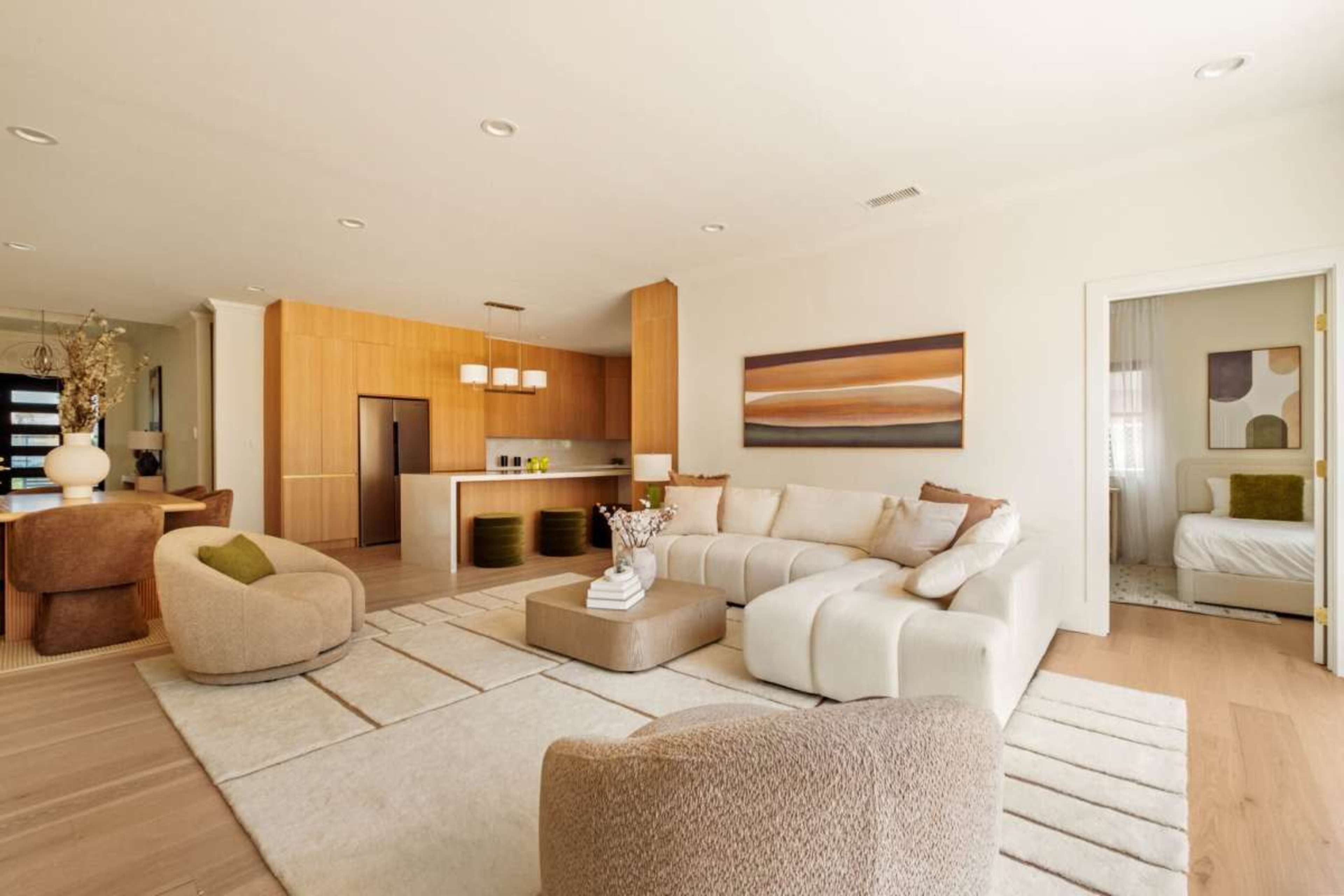 The image shows a modern living room featuring a large sectional sofa, a round accent chair, a neutral area rug, and an open kitchen area with wooden cabinetry in the background.