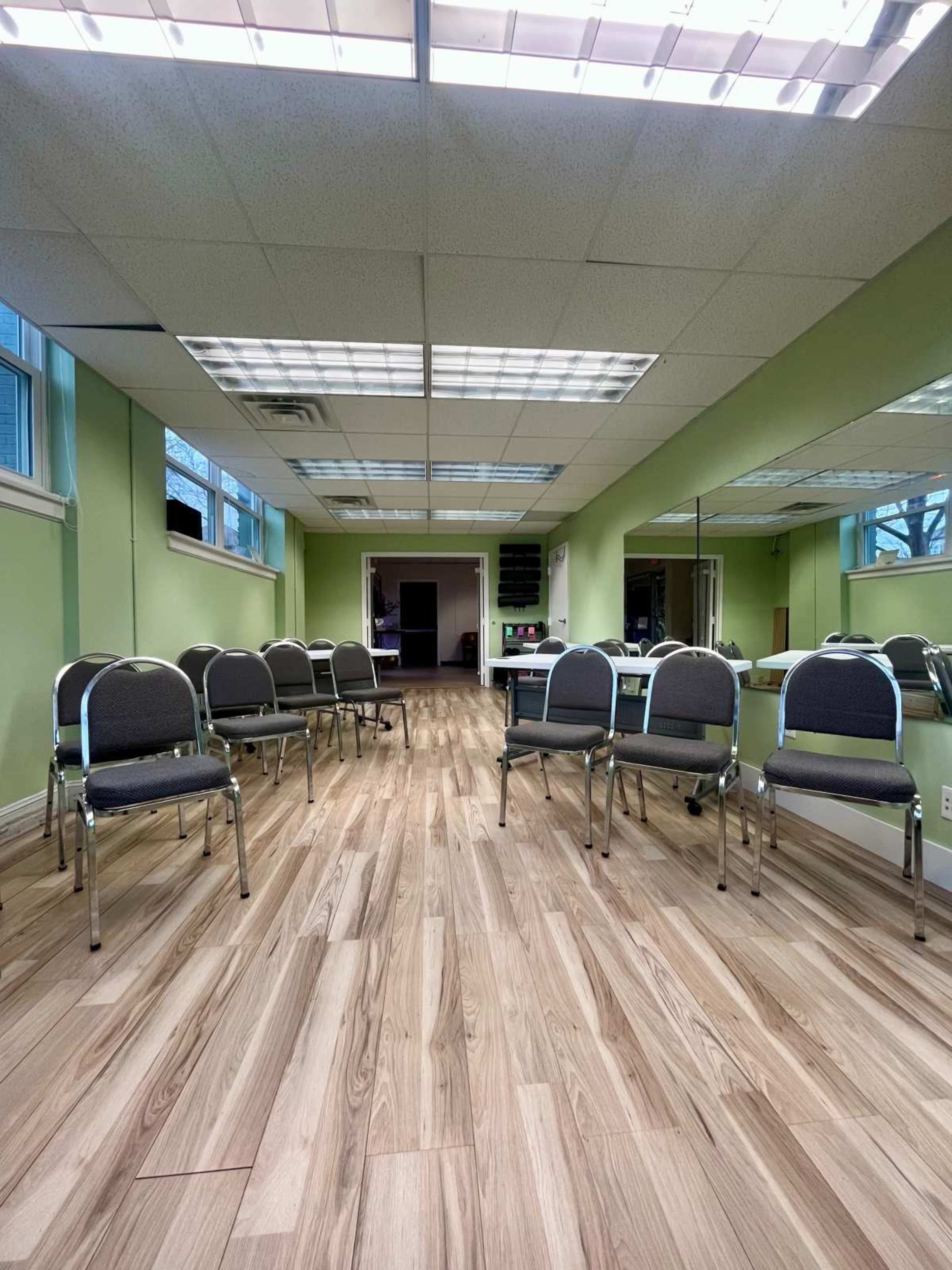 The image shows a spacious room with rows of gray chairs arranged on a light wooden floor, featuring green walls and large windows.