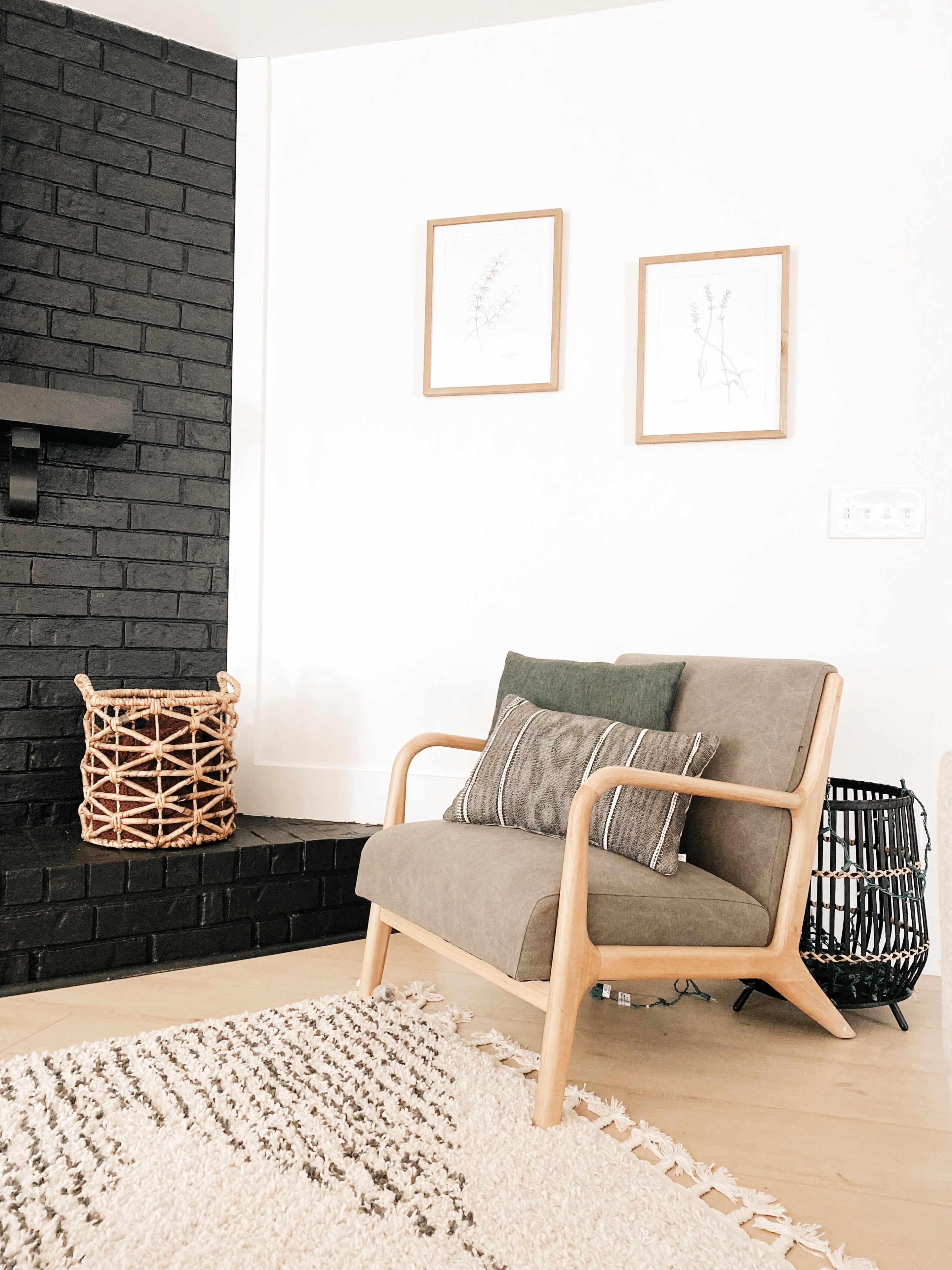 A modern living space features a light-colored chair with decorative pillows beside a black brick fireplace, complemented by framed botanical prints and woven baskets.