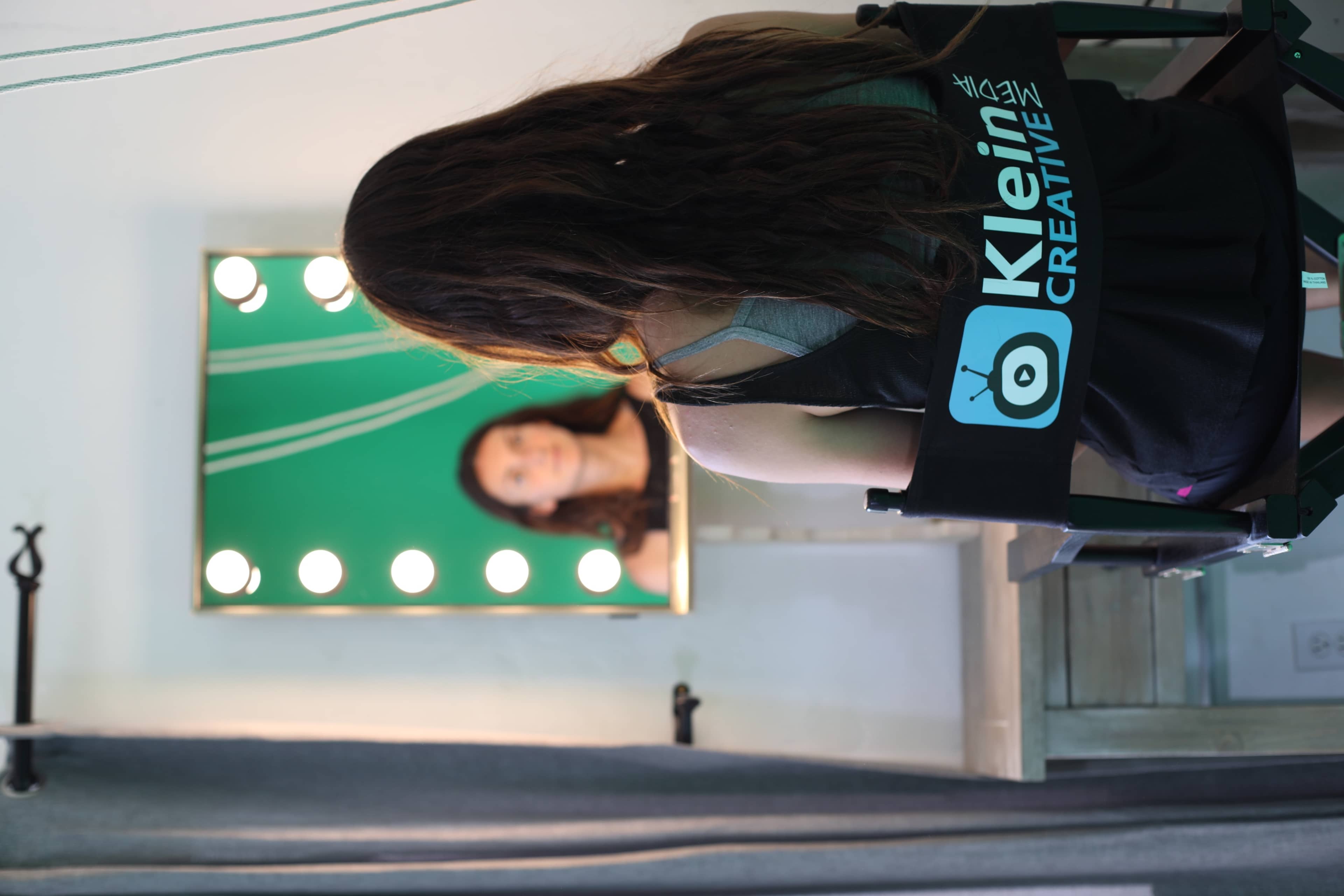 A person sits in a director's chair with "Klein Creative" on the back, facing a mirror surrounded by bright lights.