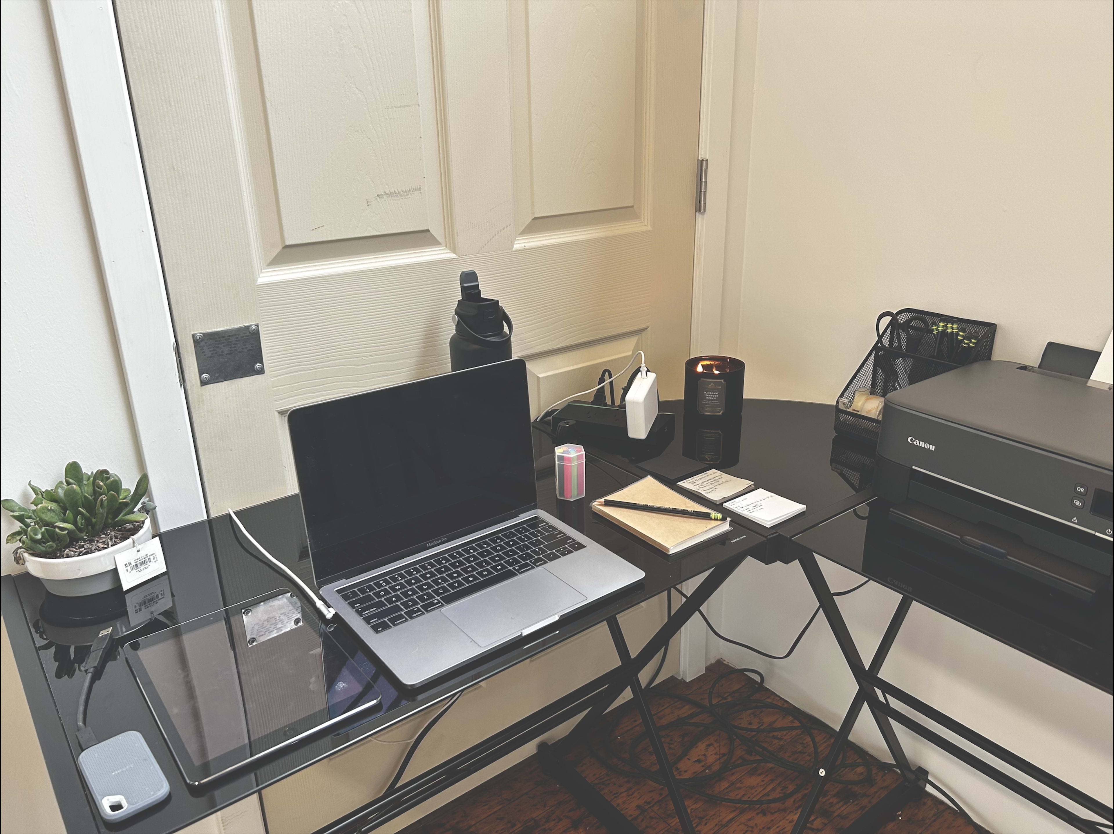 A black glass desk holds a laptop, tablet, printer, water bottle, candle, and various stationery items against a white door.