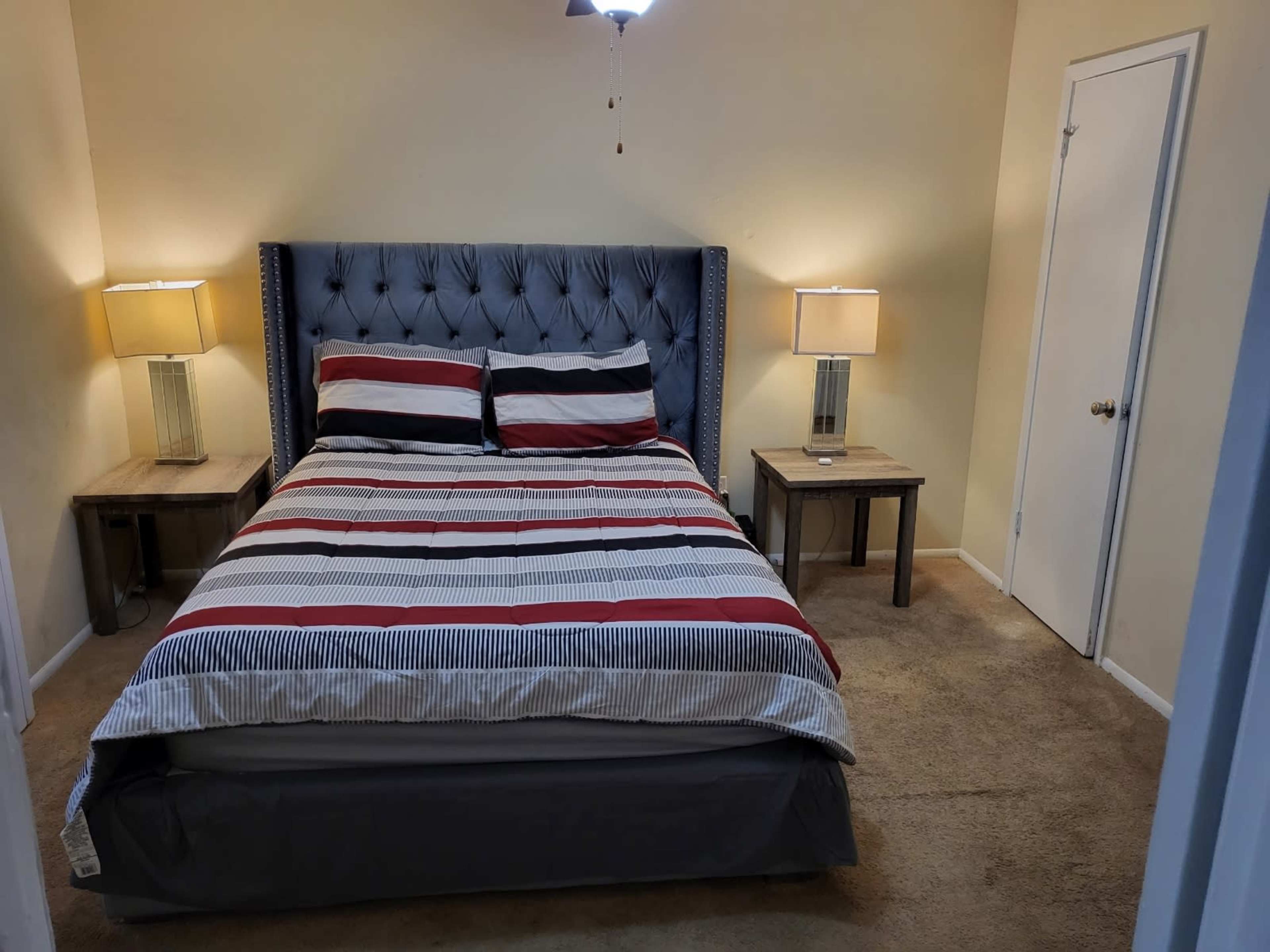 A neatly arranged bedroom features a bed with striped bedding, two side tables with lamps, and a door leading to another room.