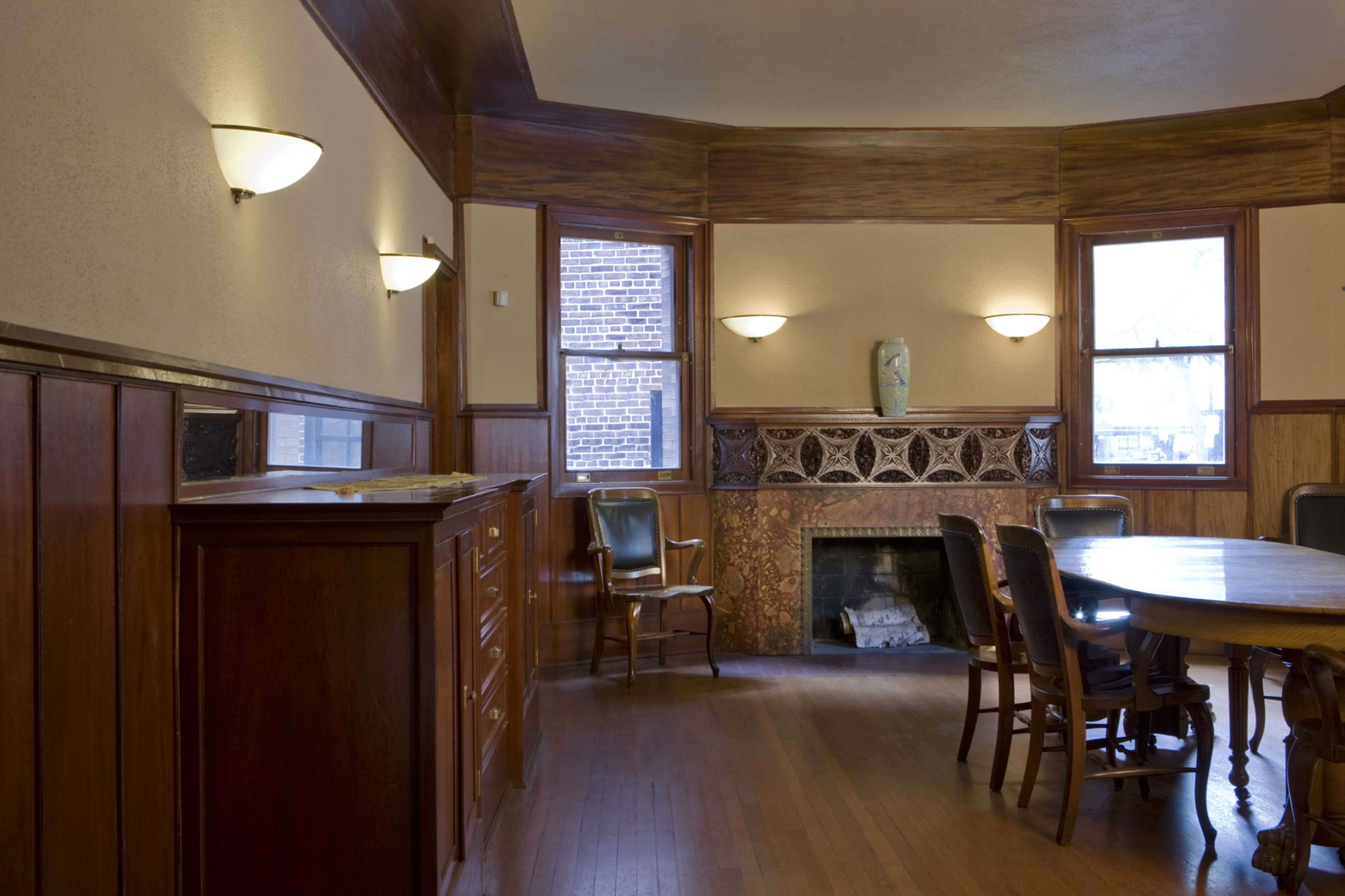 A wooden-paneled room with a round table surrounded by chairs, a fireplace with decorative tiles, and windows with light fixtures on the walls.
