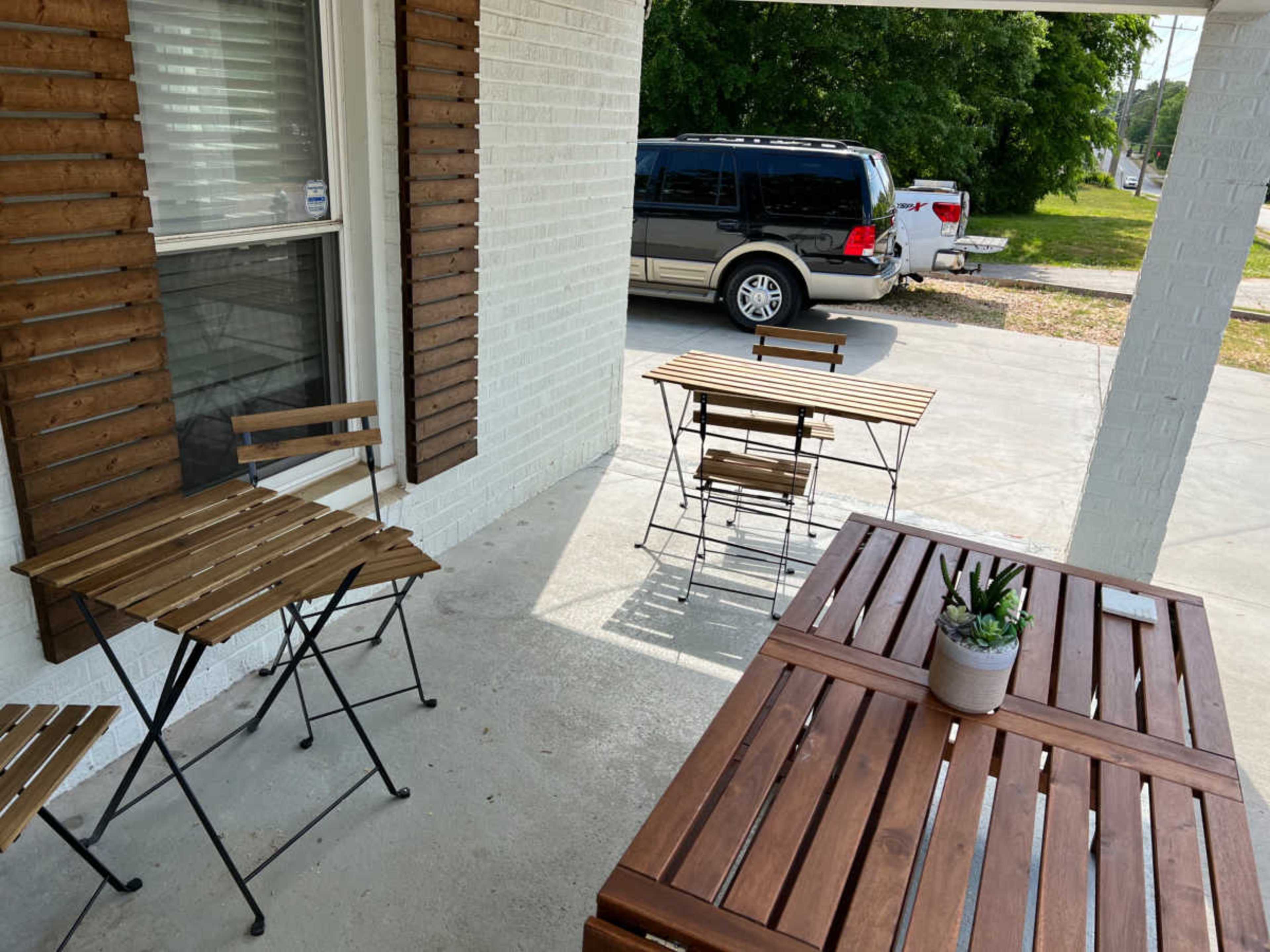 The image shows a porch area with two small wooden tables and chairs, along with a larger wooden table, while a parked SUV is visible in the background.