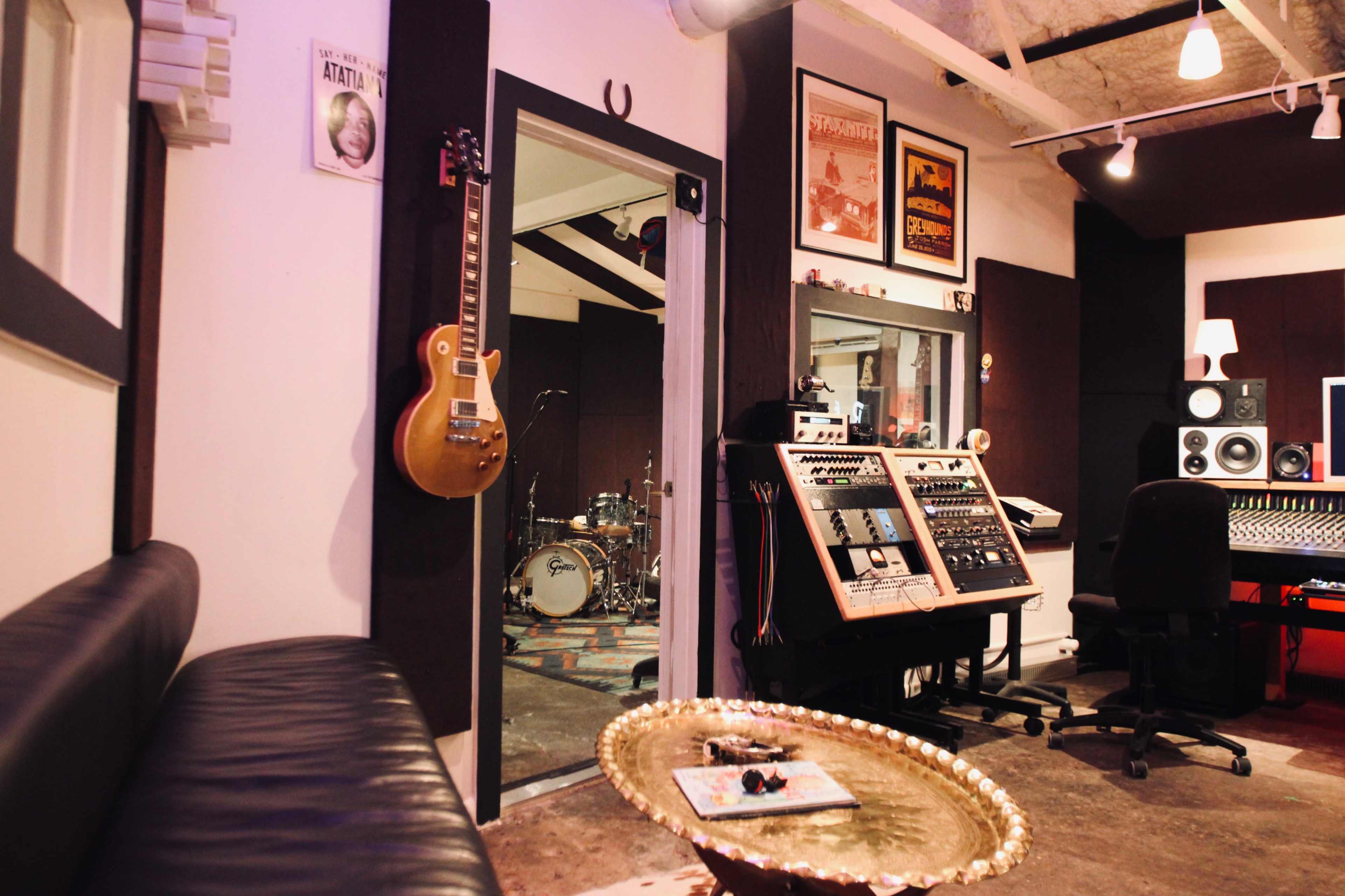 The image shows a music recording studio with a guitar on the wall, a control panel, and a visible drum set in a separate room.