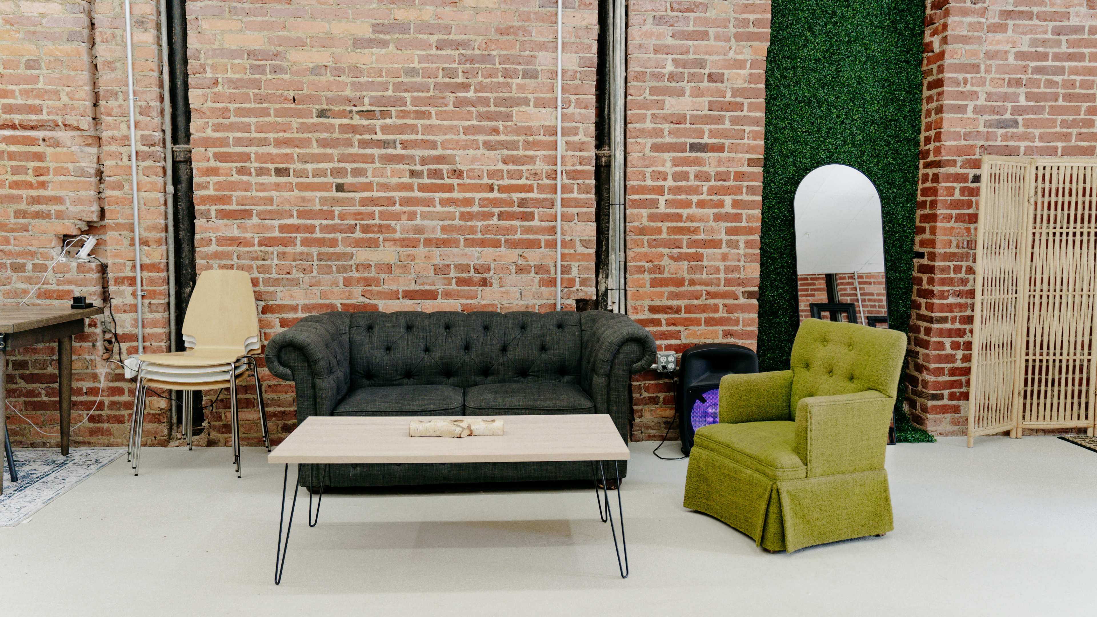 The image shows a modern interior with a black tufted sofa, a green upholstered armchair, a wooden coffee table, and exposed brick walls.