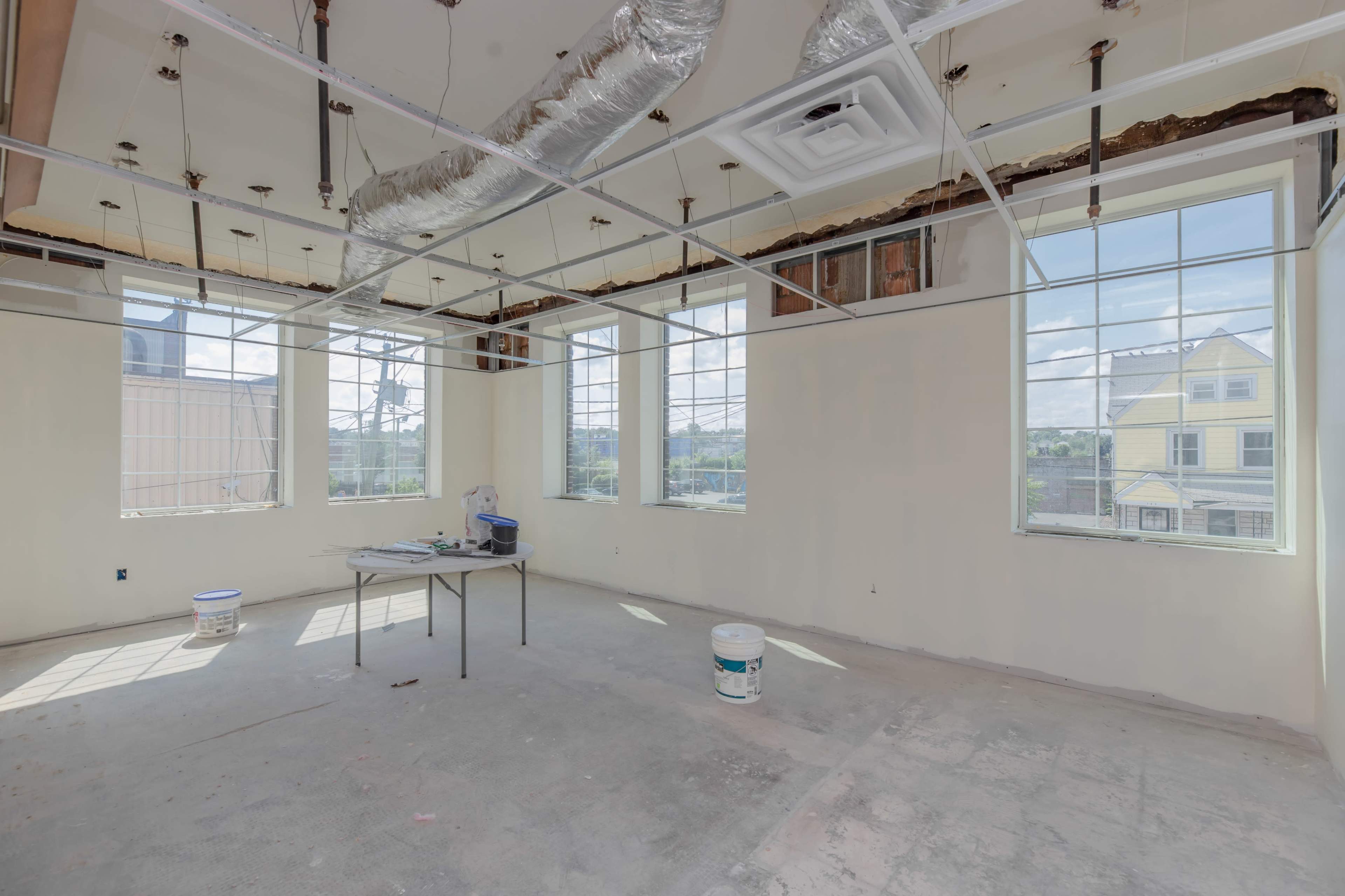 An empty room with unfinished walls, exposed ceiling structures, and large windows allowing natural light to enter.