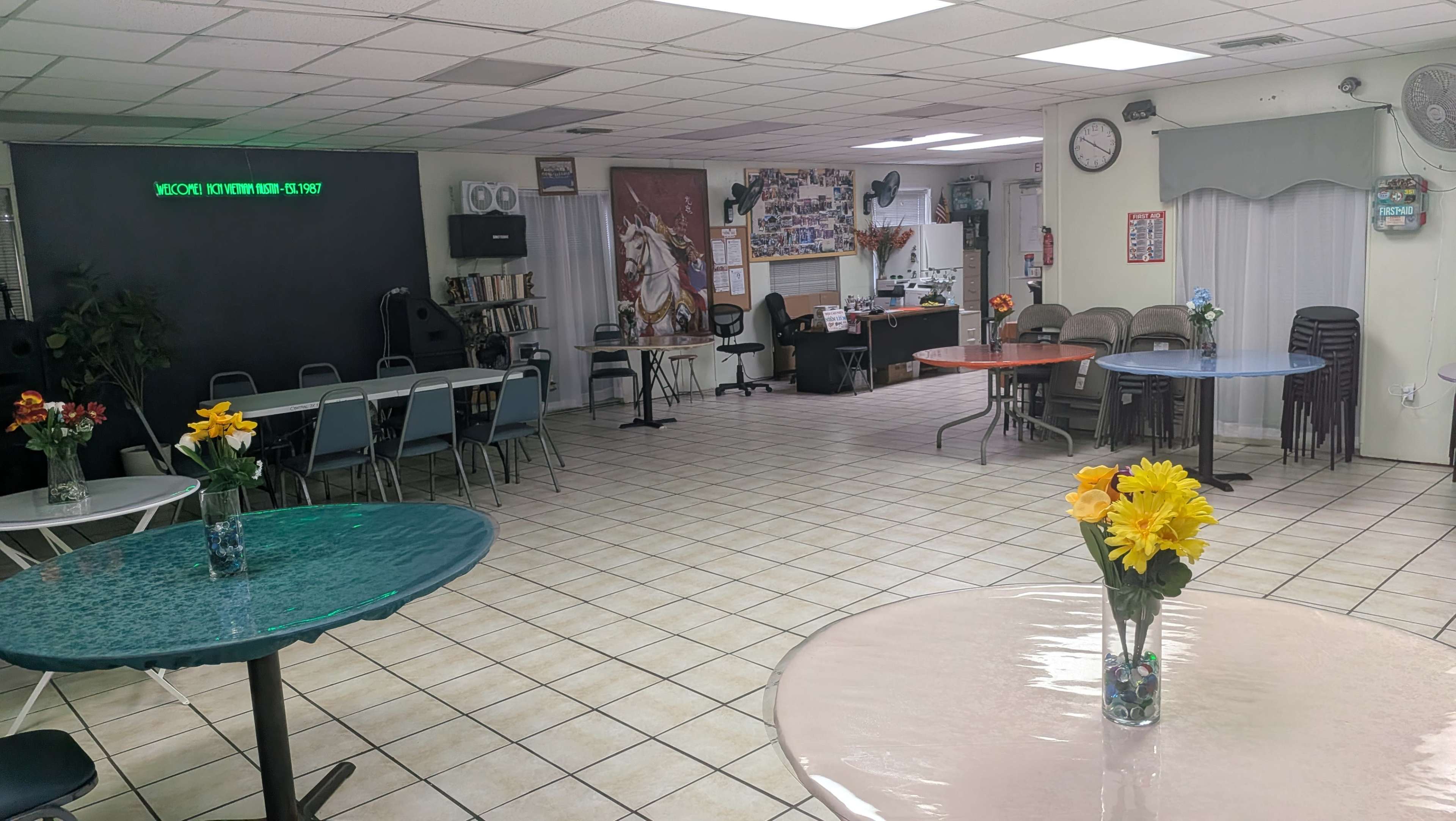 The image shows an empty community room with several tables set with vases of flowers and chairs arranged around them, while various decorations and a service area can be seen in the background.
