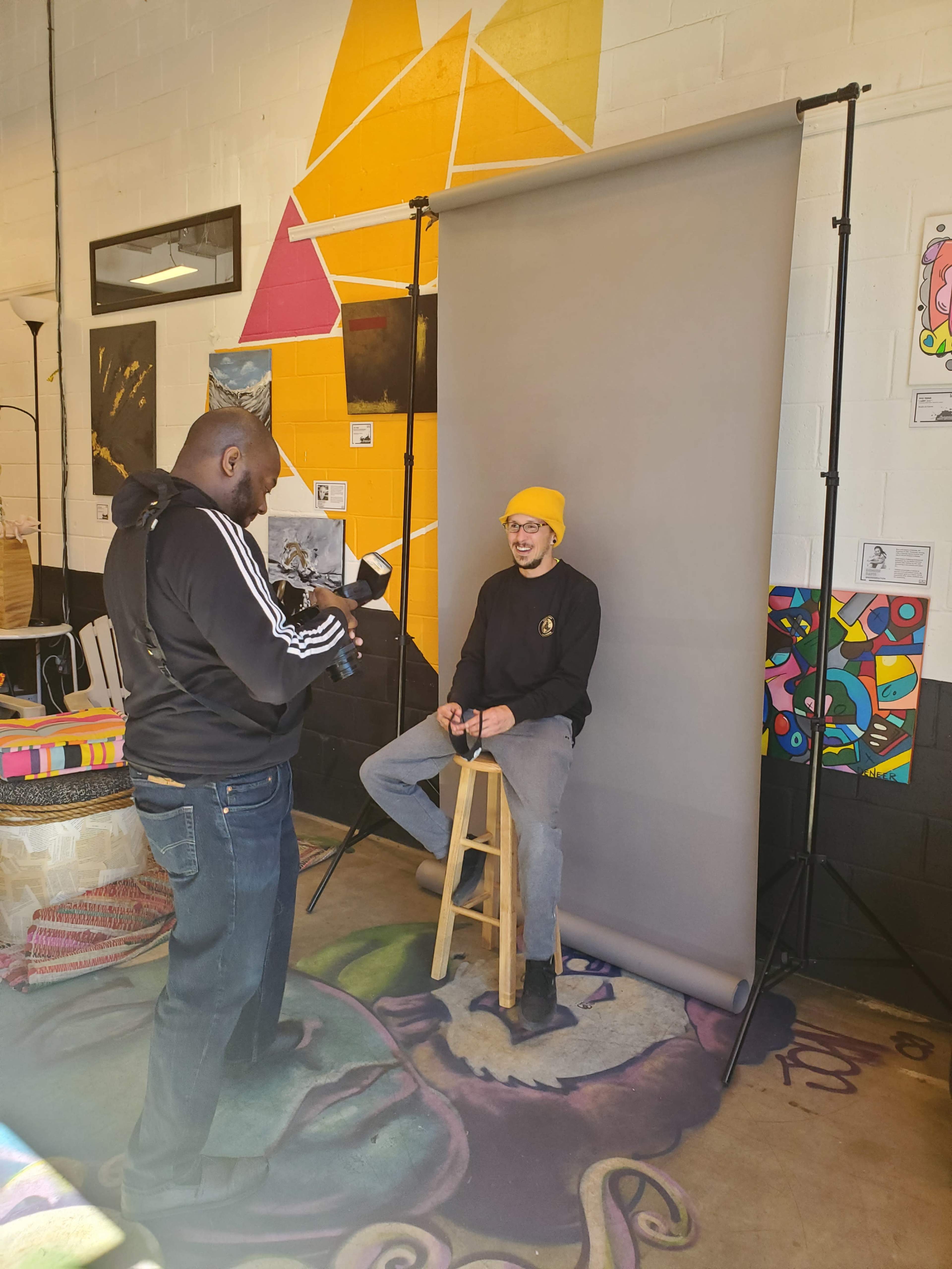 A photographer is capturing an image of a person sitting on a stool in front of a gray backdrop in an art-filled studio.