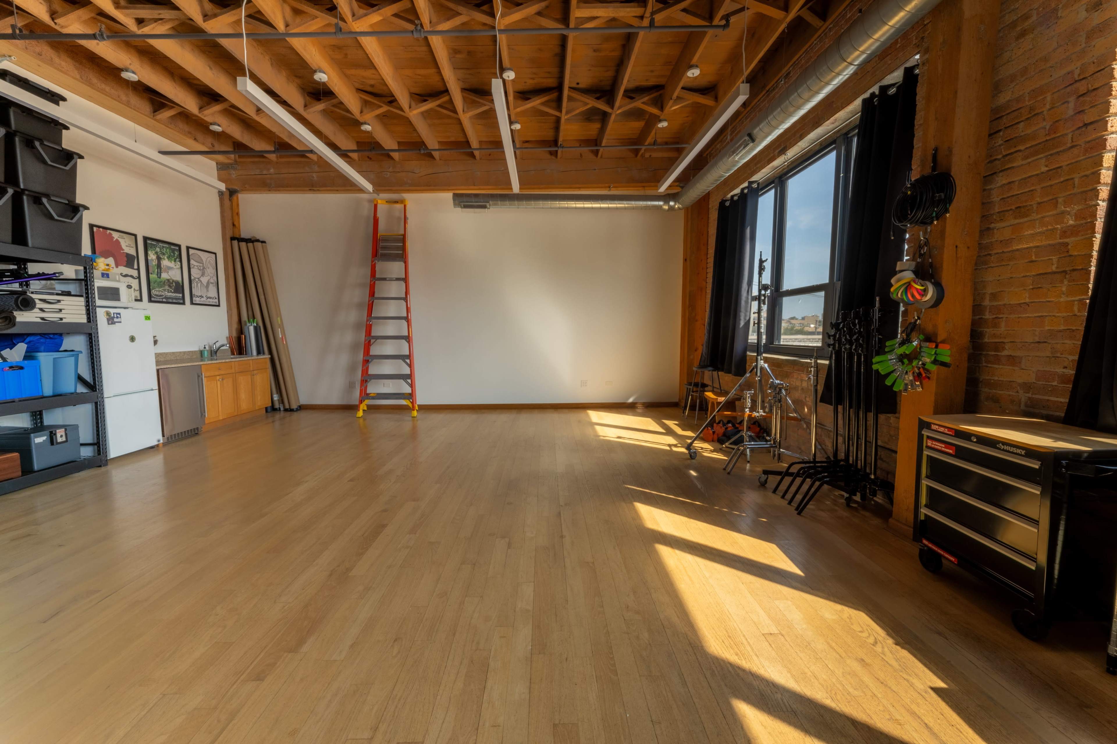An empty room with hardwood floors, exposed wooden beams, and large windows, featuring a ladder, storage boxes, and various tools against the walls.