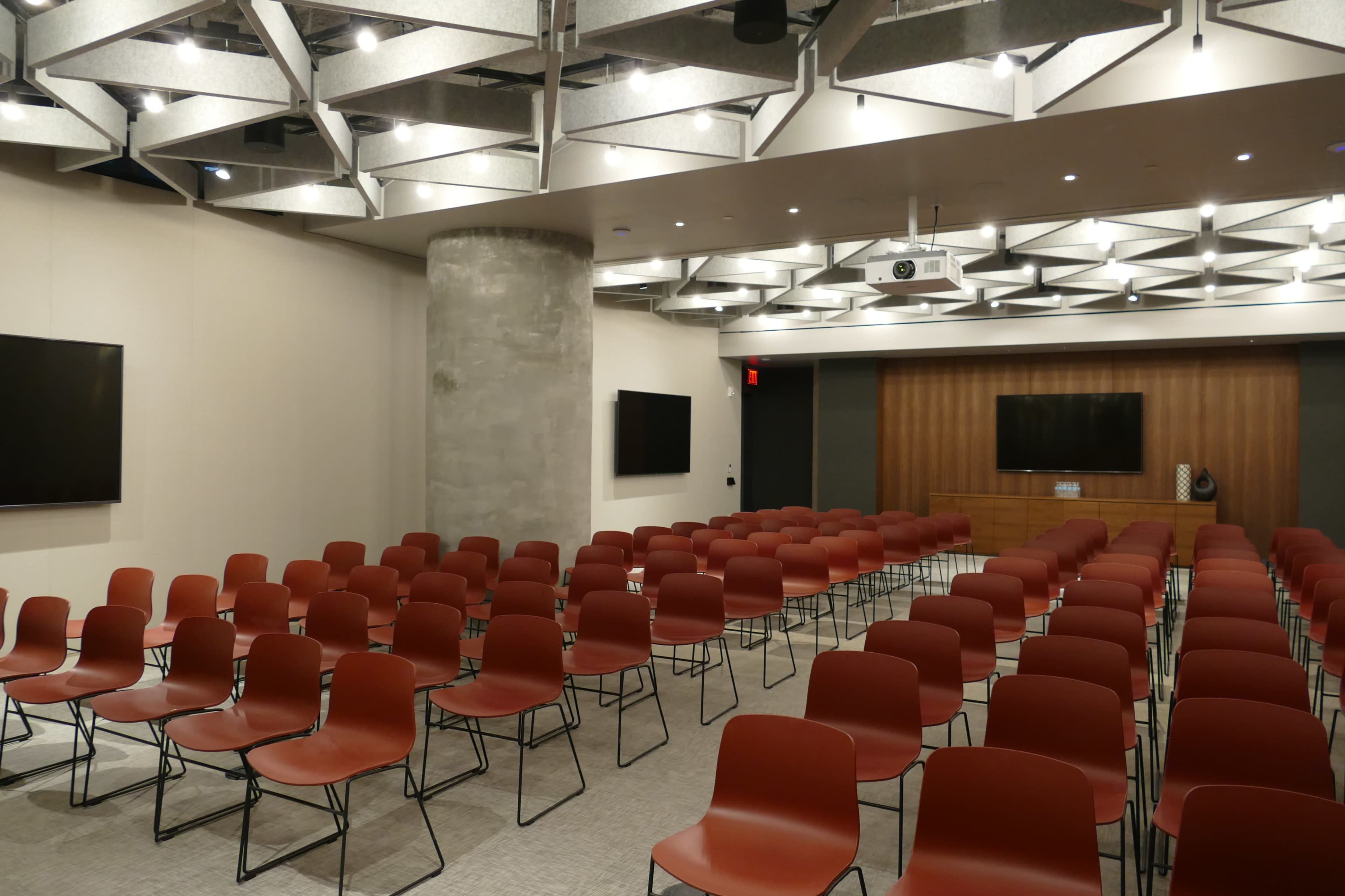 A modern conference room set up with multiple rows of red chairs facing a wooden presentation area, featuring two large screens on the walls.