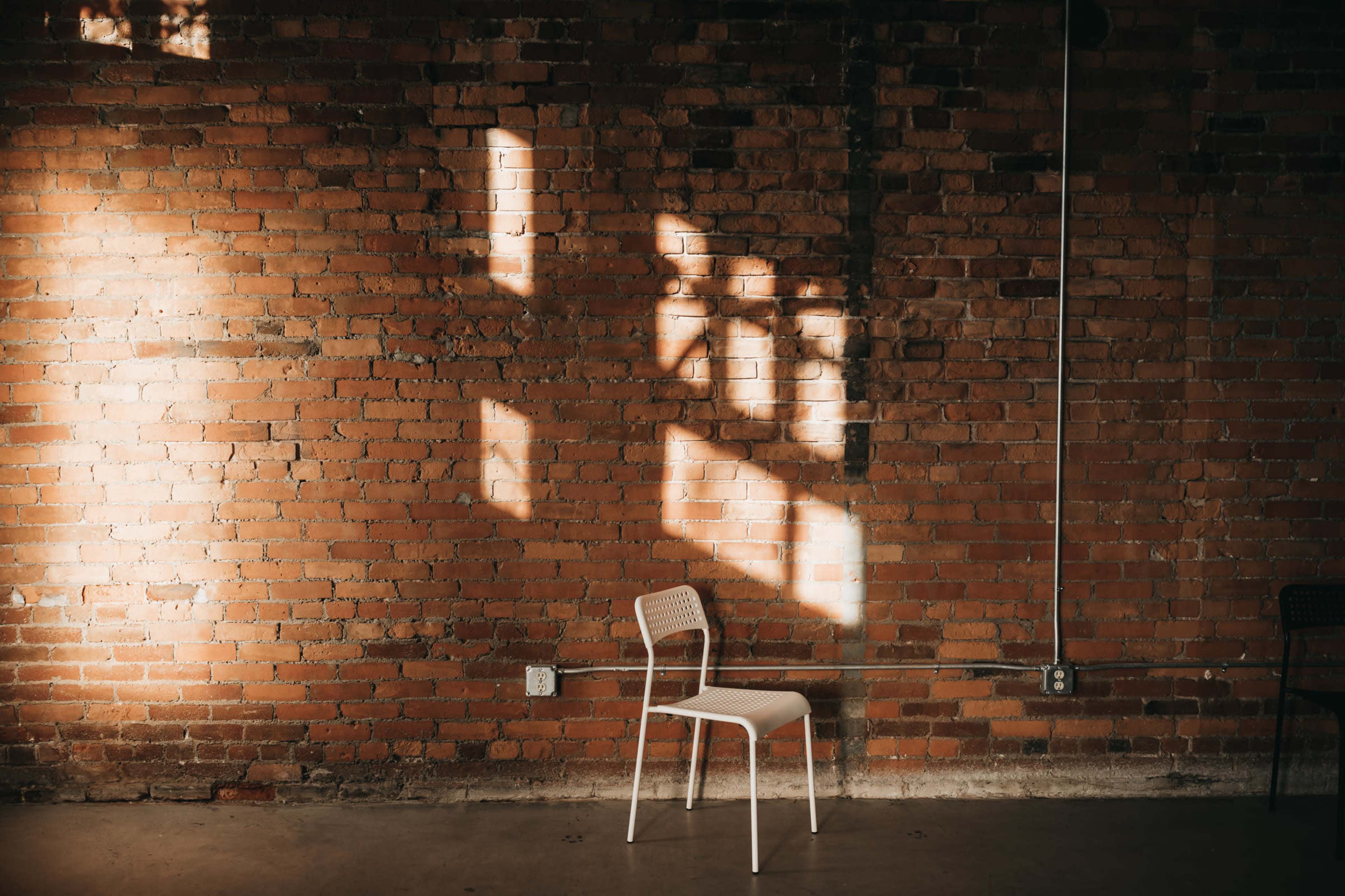 A white chair sits against a brick wall illuminated by sunlight casting shadows.